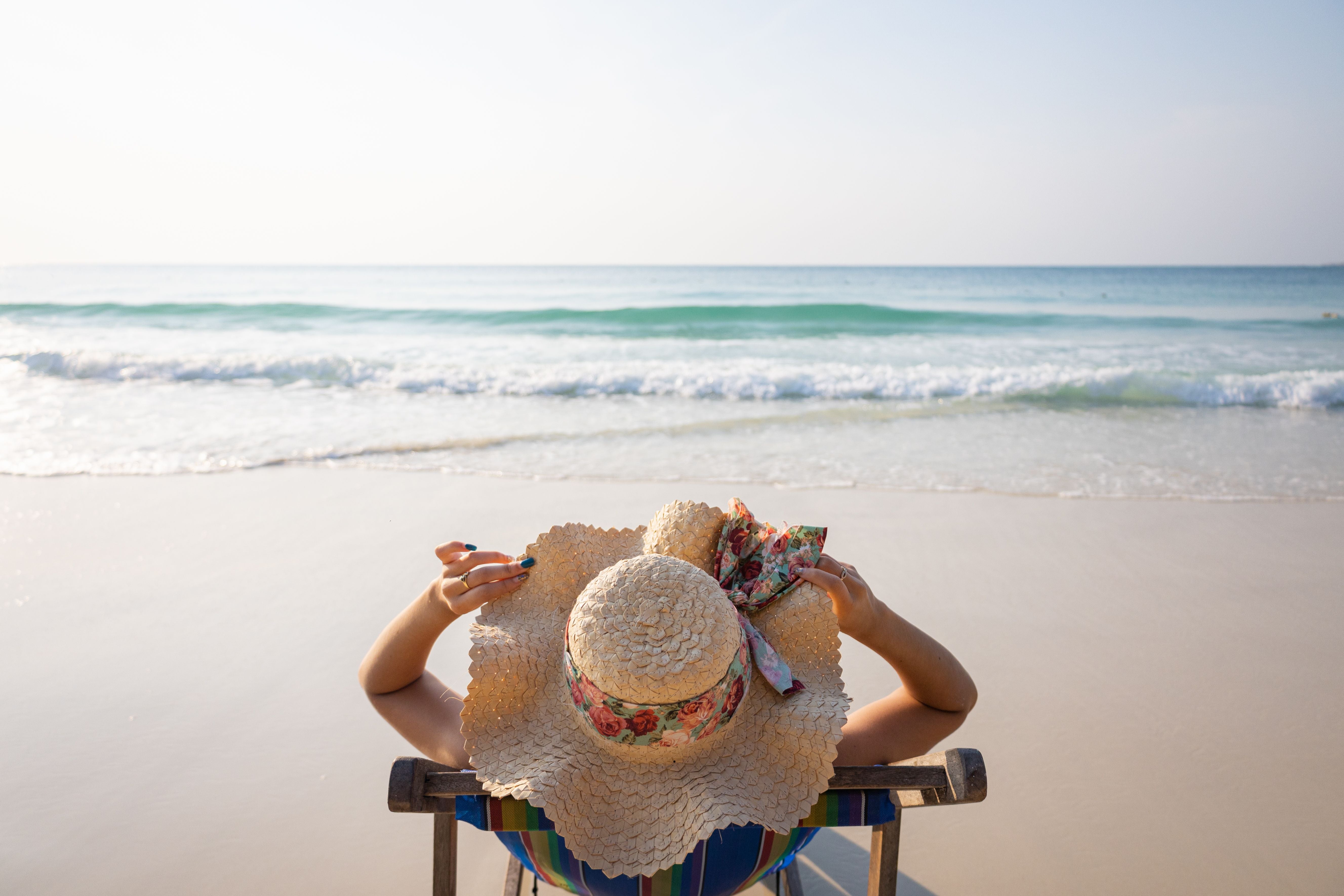 Frau mit Sonnenhut entspannt auf einem Liegestuhl am Strand mit Blick aufs Meer.
