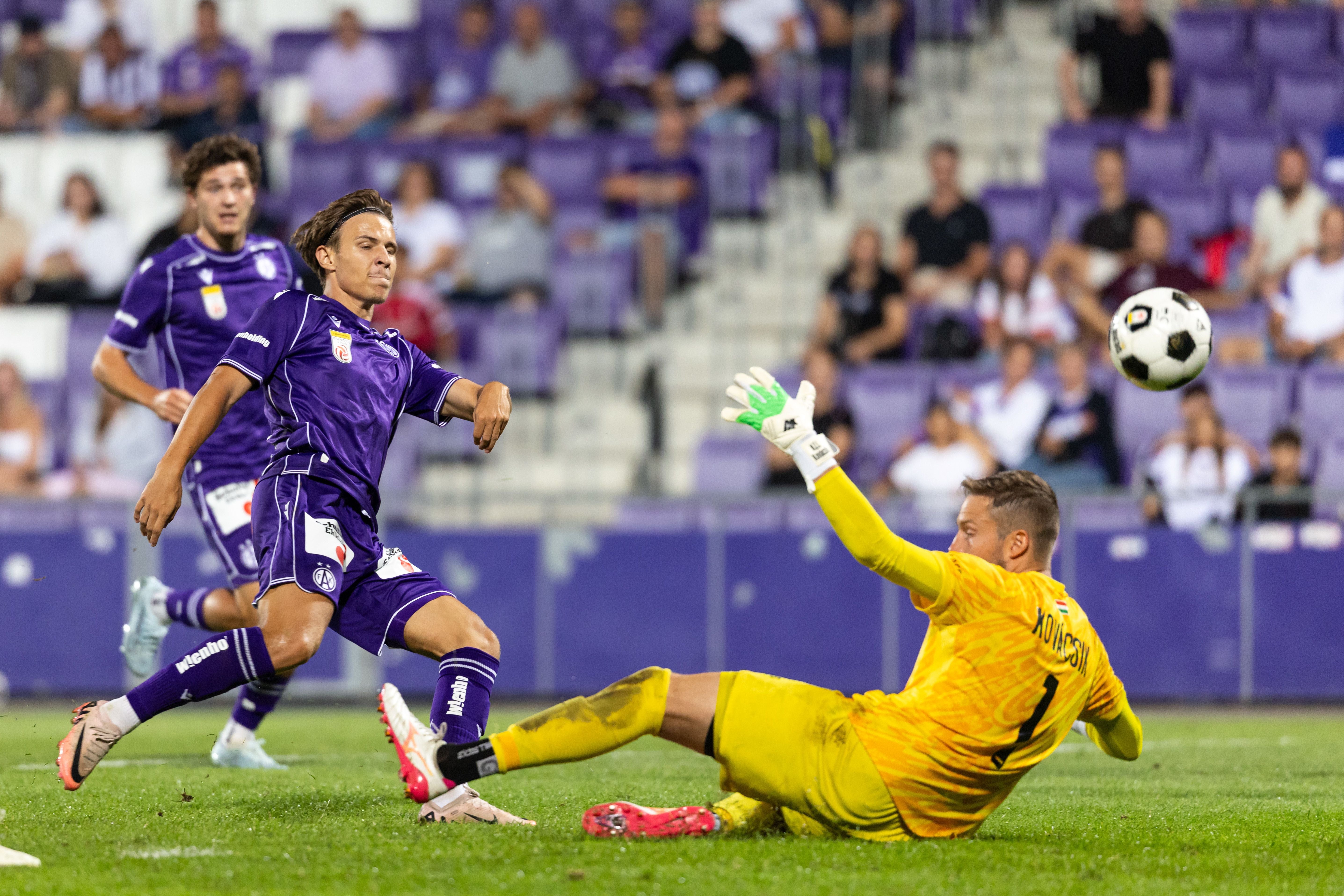 Ein Fußballspieler in einem violetten Trikot schießt im Lauf den Ball am am Boden liegenden Torwart in gelber Kleidung vorbei. Im Hintergrund sind besetzte Tribünen in einem Fußballstadion erkennbar.