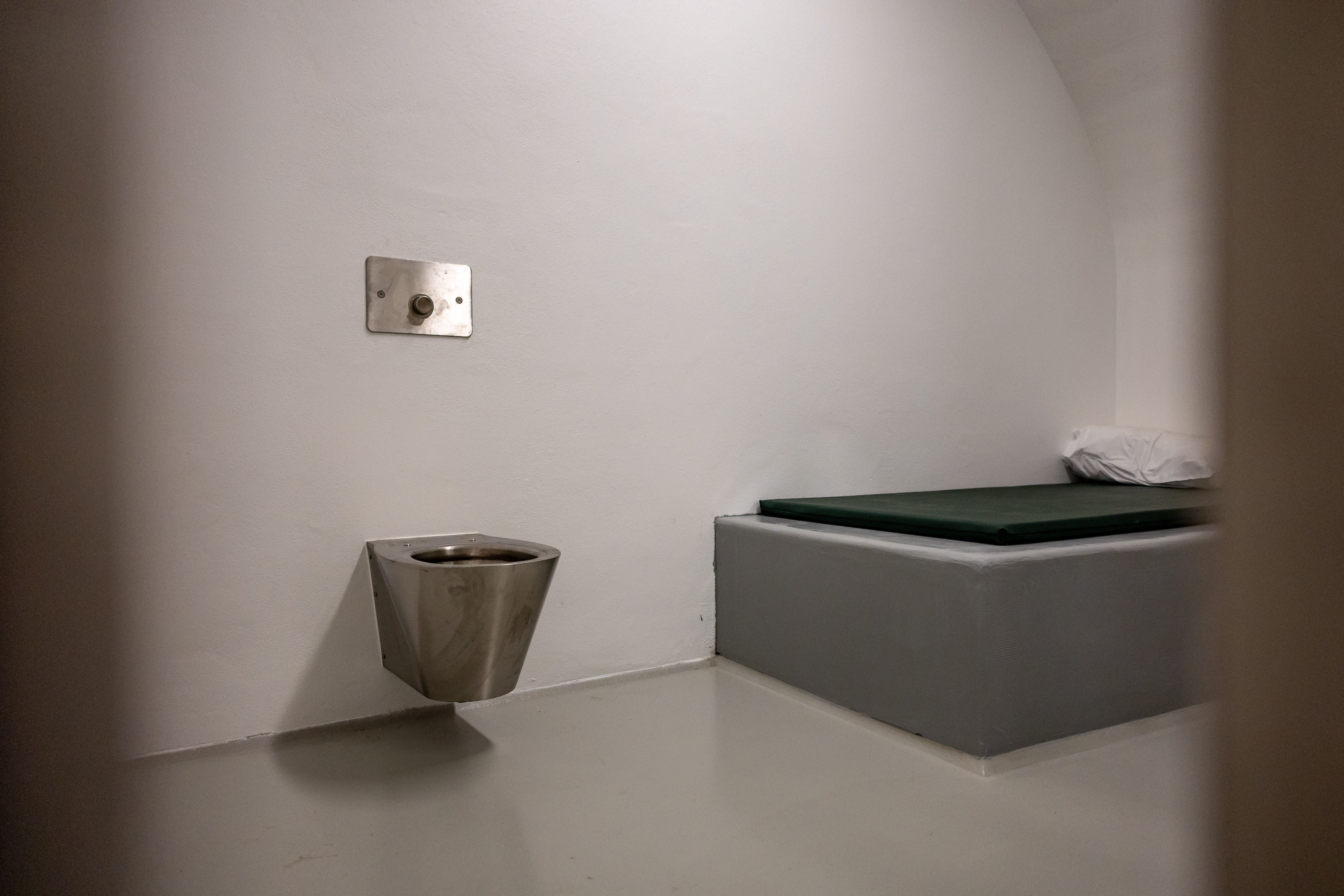 Kleine, schlichte Gefängniszelle mit Metalltoilette und Pritsche mit Matratze und Kissen.