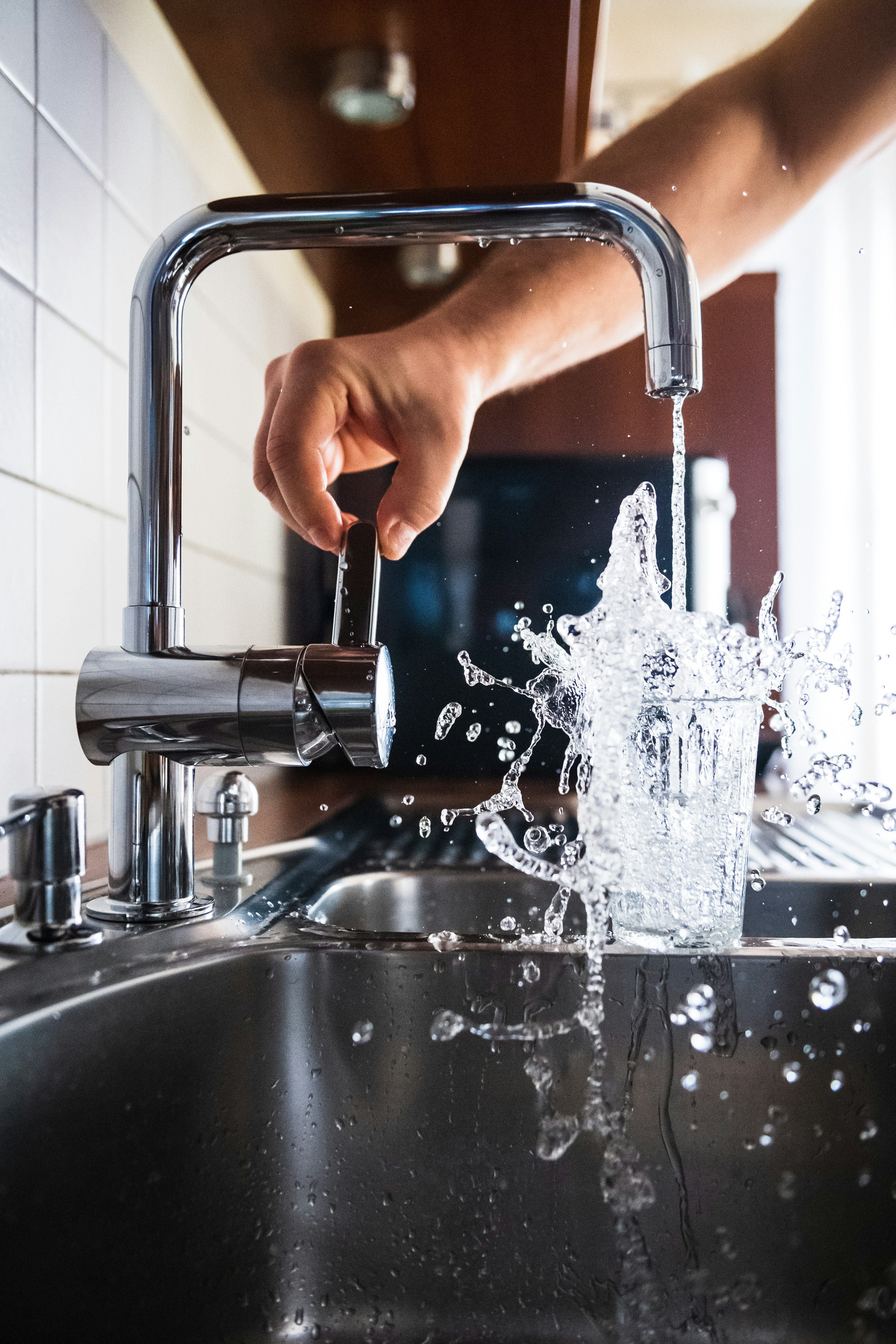 Wasserglas unter laufendem Wasserhahn, aus dem Glas spritzt Wasser über das Spülbecken.