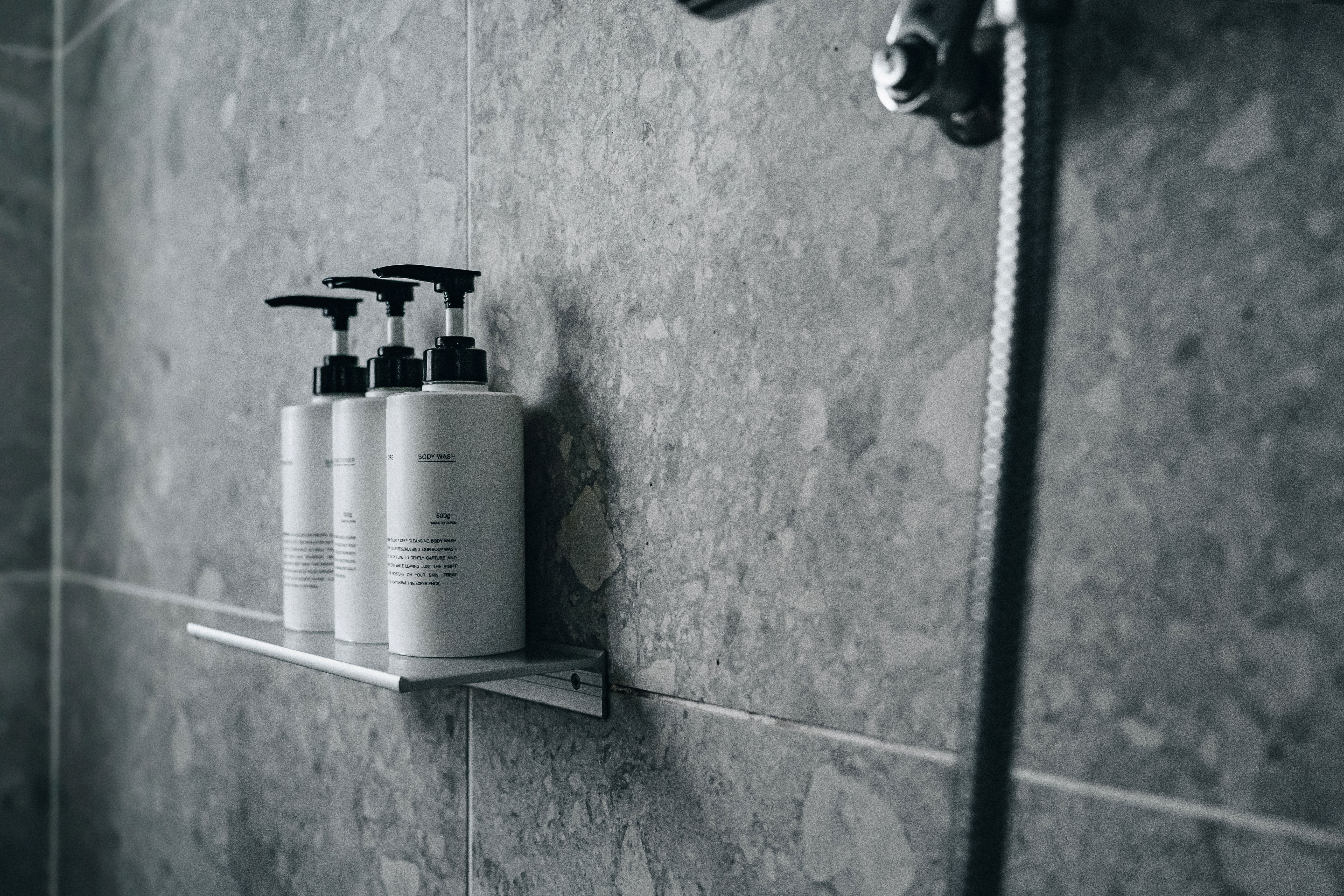 Drei Spenderflaschen auf Metallablage vor grauen Fliesen in einer modernen Dusche.