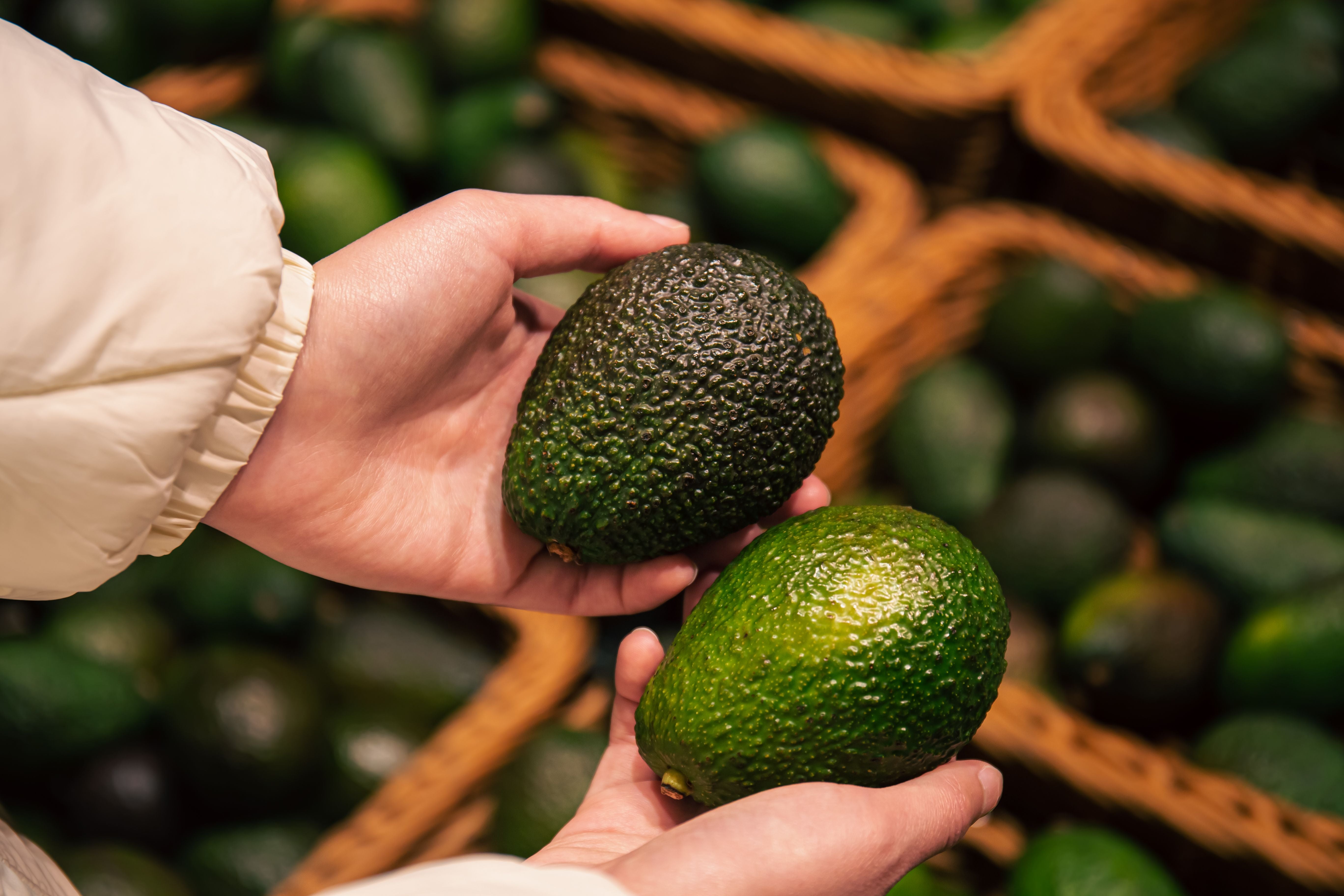 Zwei Avocados werden in den Händen einer Person im Supermarkt gehalten und verglichen.