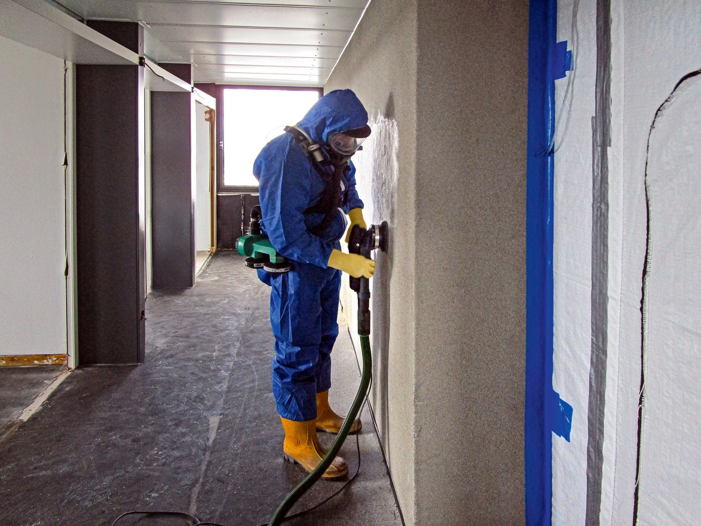 Person in Schutzanzug schleift eine Wand, vermutlich bei einer Schadstoffsanierung.