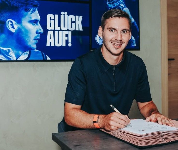 Ein Mann unterschreibt einen Vertrag, im Hintergrund steht "Glück auf!" und Schalke 04 Logo ist sichtbar.