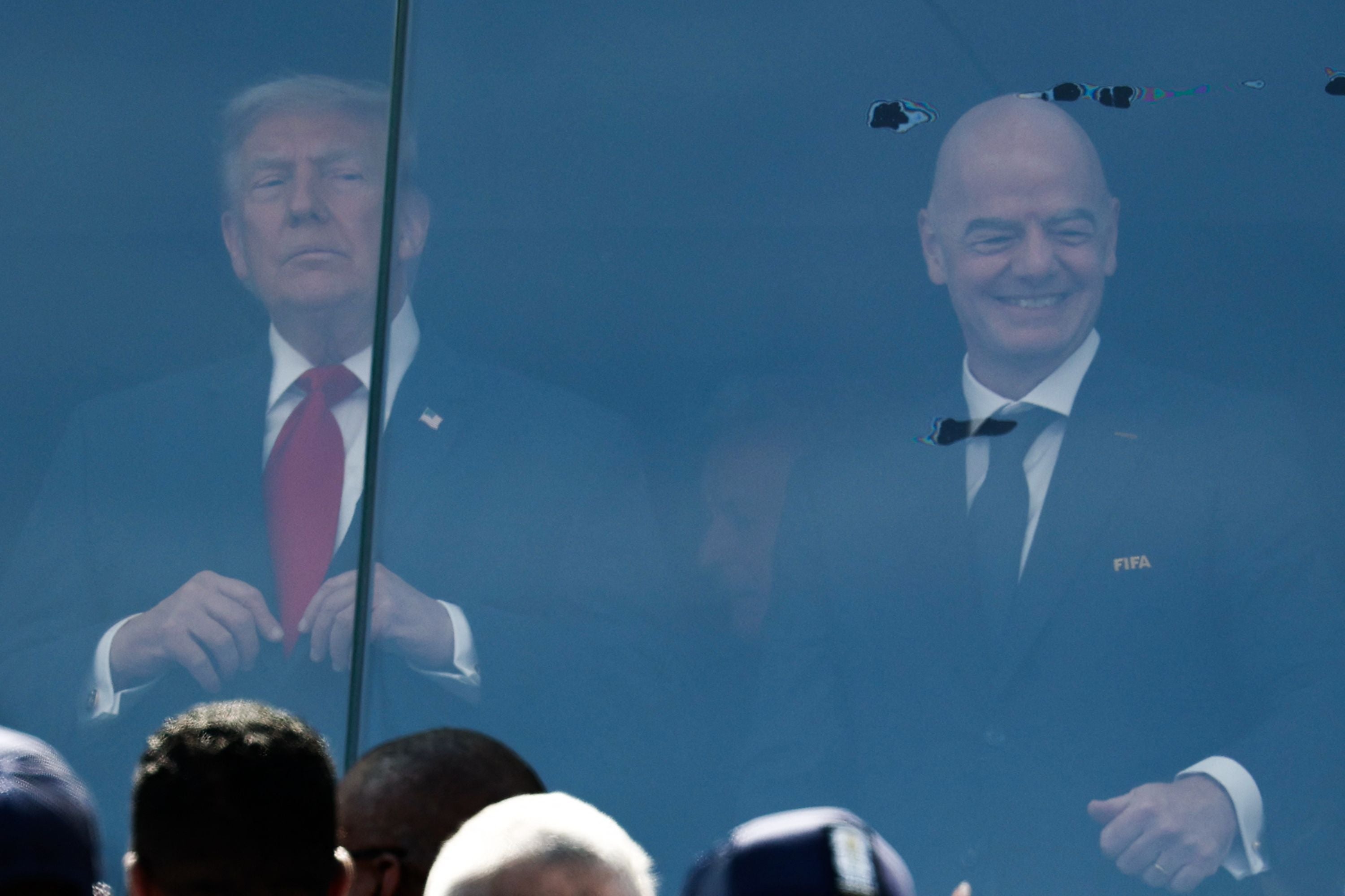 Zwei Männer im Anzug stehen hinter einer Glasscheibe im Stadion und beobachten ein Fußballspiel.