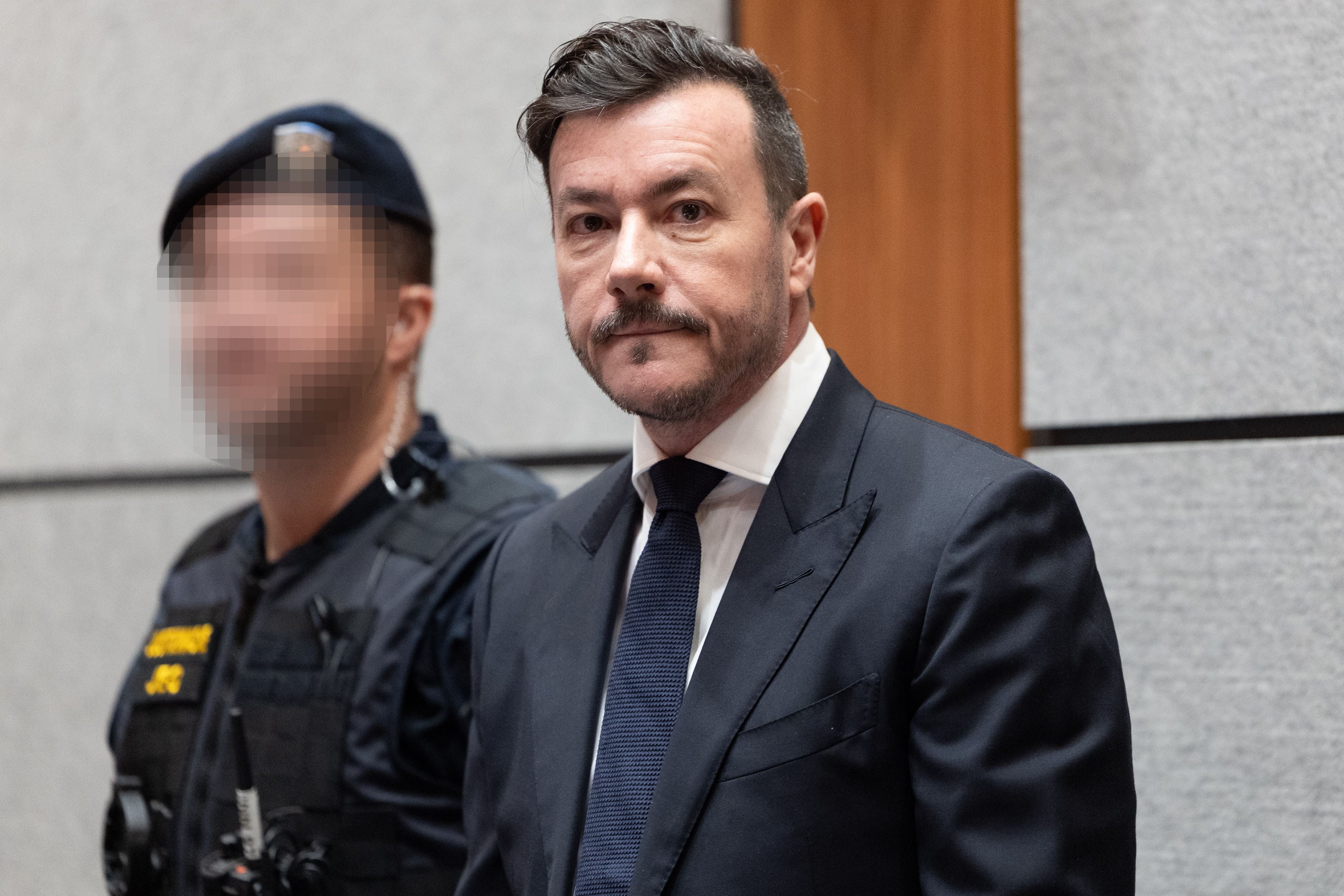 Ein Mann im Anzug steht in einem Gerichtssaal neben einem unscharf dargestellten Polizisten.