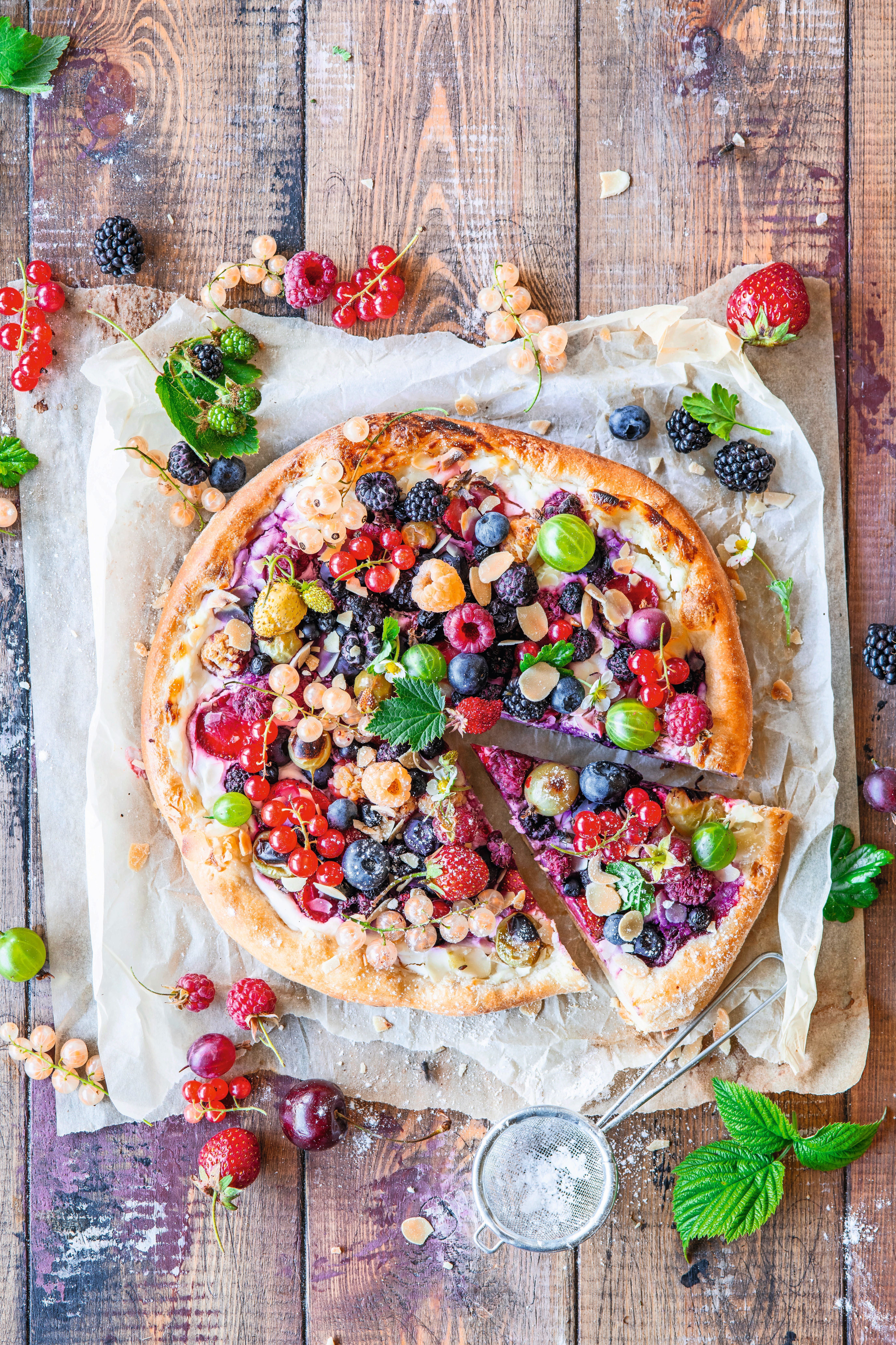 Bunte Pizza mit verschiedenen frischen Beeren auf Backpapier, rustikaler Holzhintergrund.