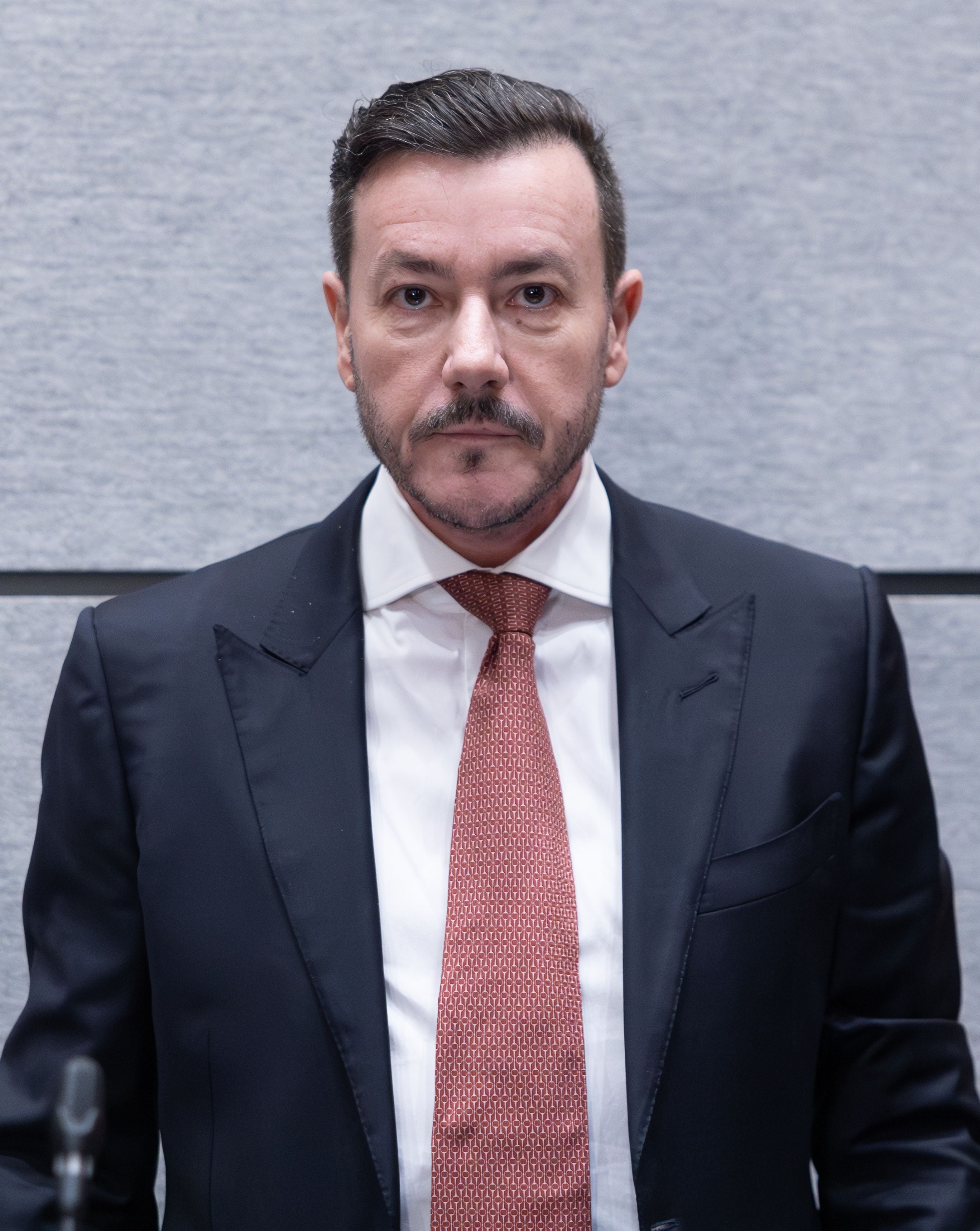 Ein Mann im Anzug mit roter Krawatte steht vor einer grauen Wand und blickt ernst in die Kamera.