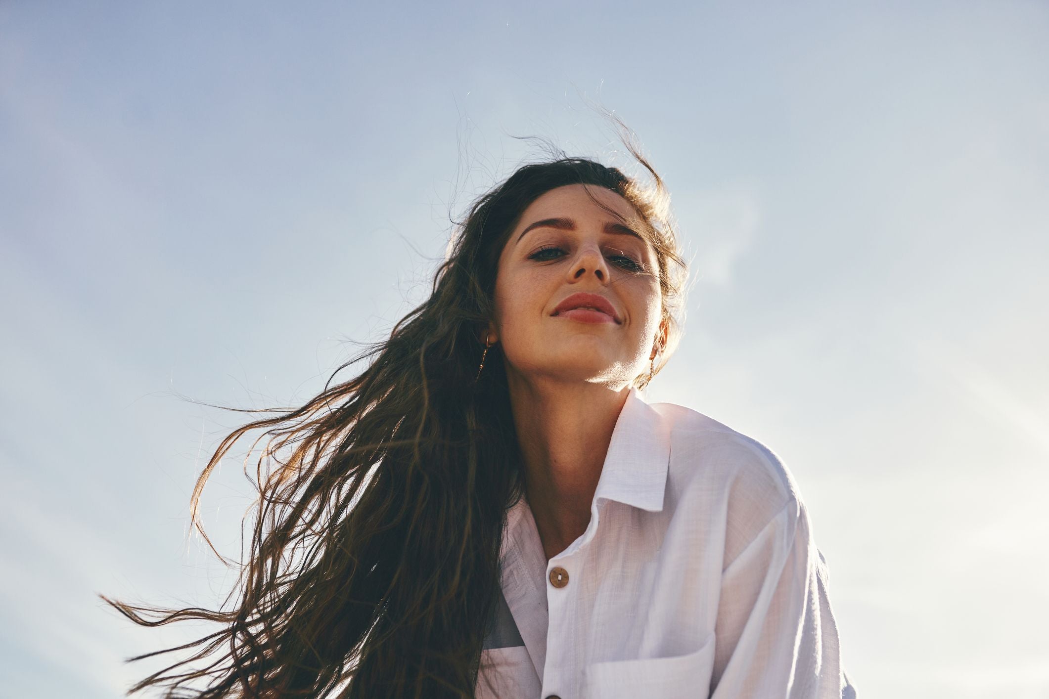 Frau mit langen, wehenden Haaren blickt hinab, Sonnenlicht und klarer Himmel im Hintergrund.