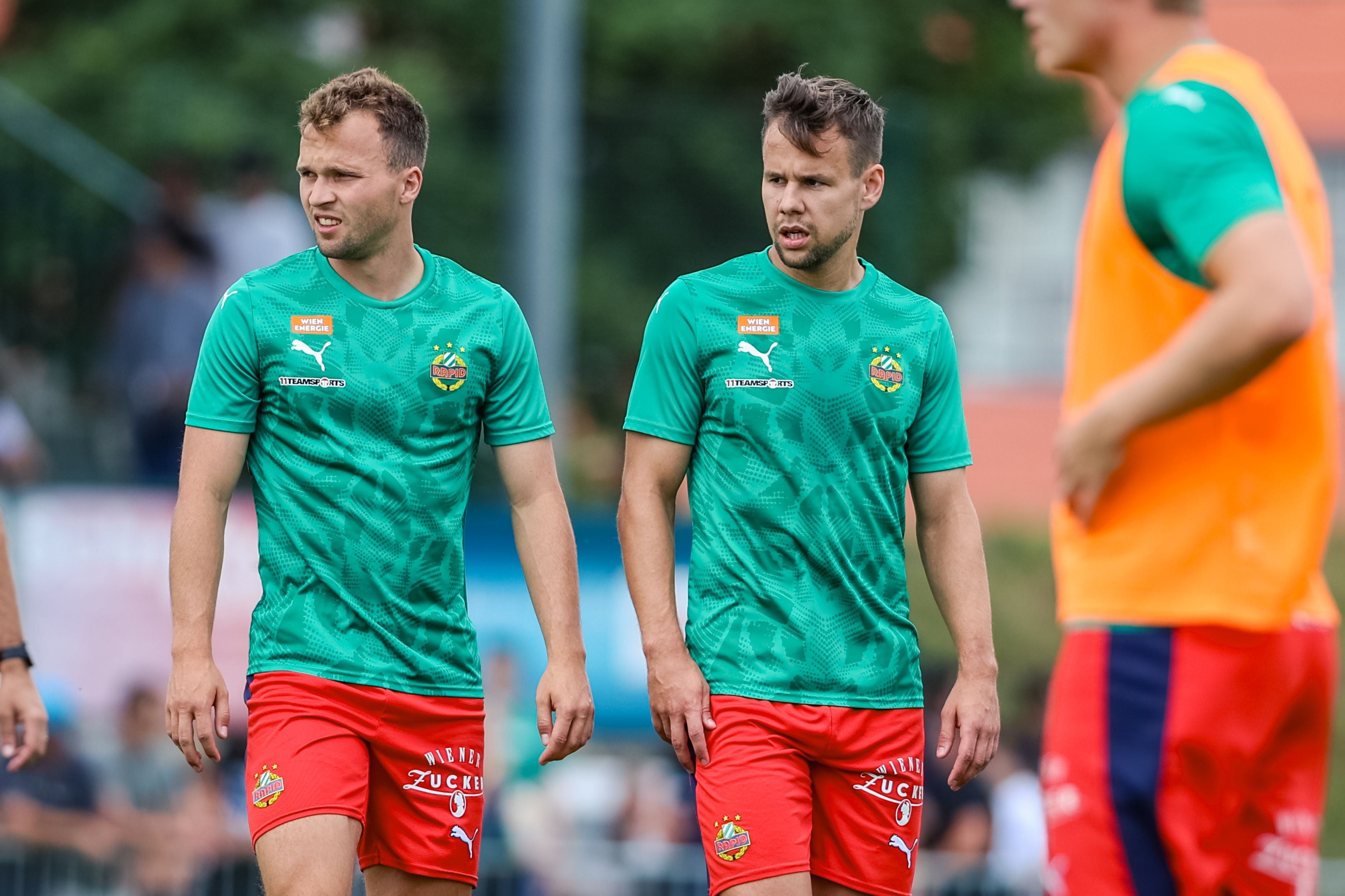 Zwei Männer in grünen Sport-T-Shirts und roten Shorts gehen nebeneinander über einen Rasenplatz. Neben ihnen ist eine weitere Person in einem orangefarbenen Trainingsleibchen zu sehen.