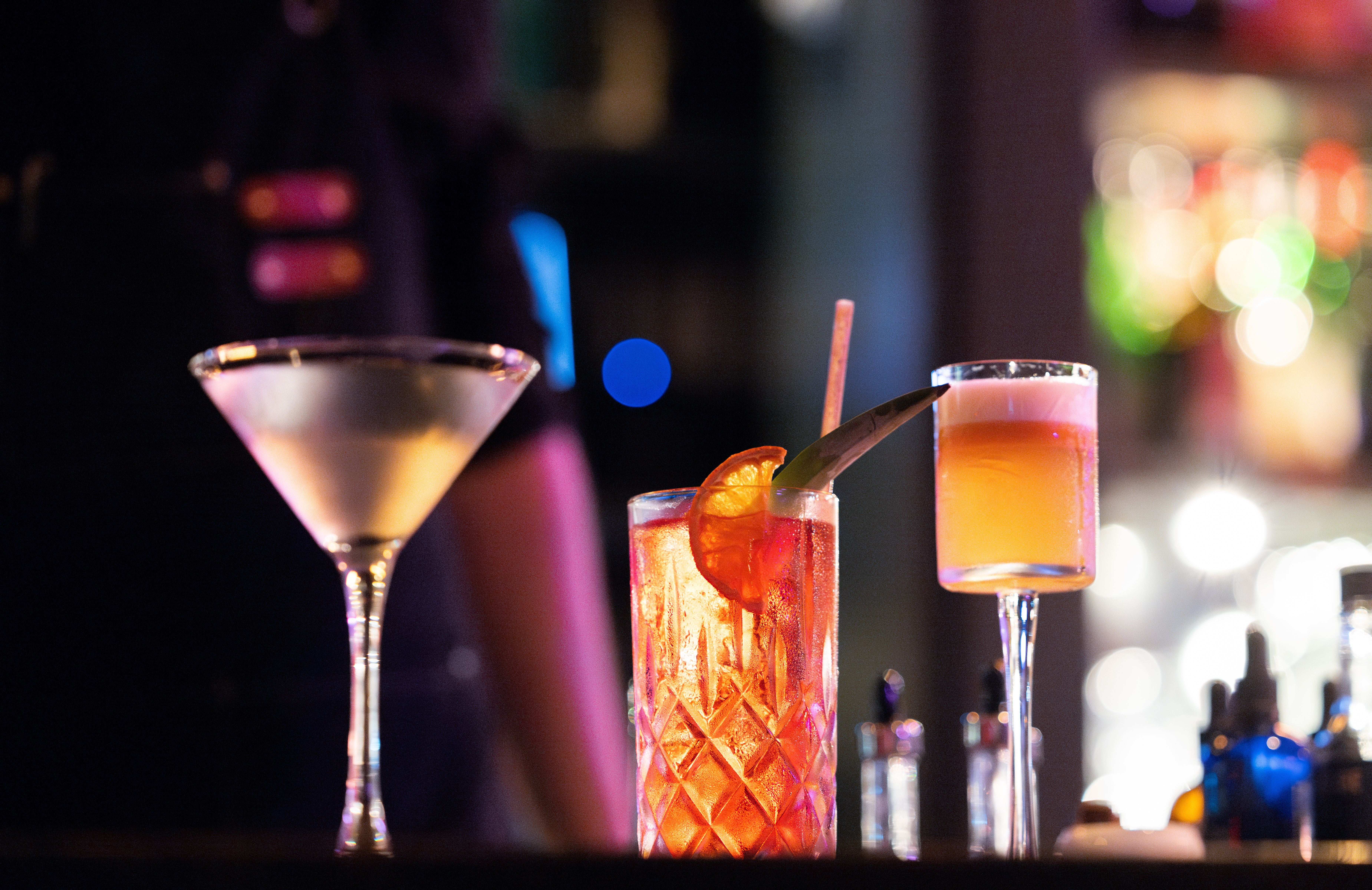 Drei Cocktails stehen beleuchtet auf einer Bar, der Hintergrund ist dunkel und verschwommen.