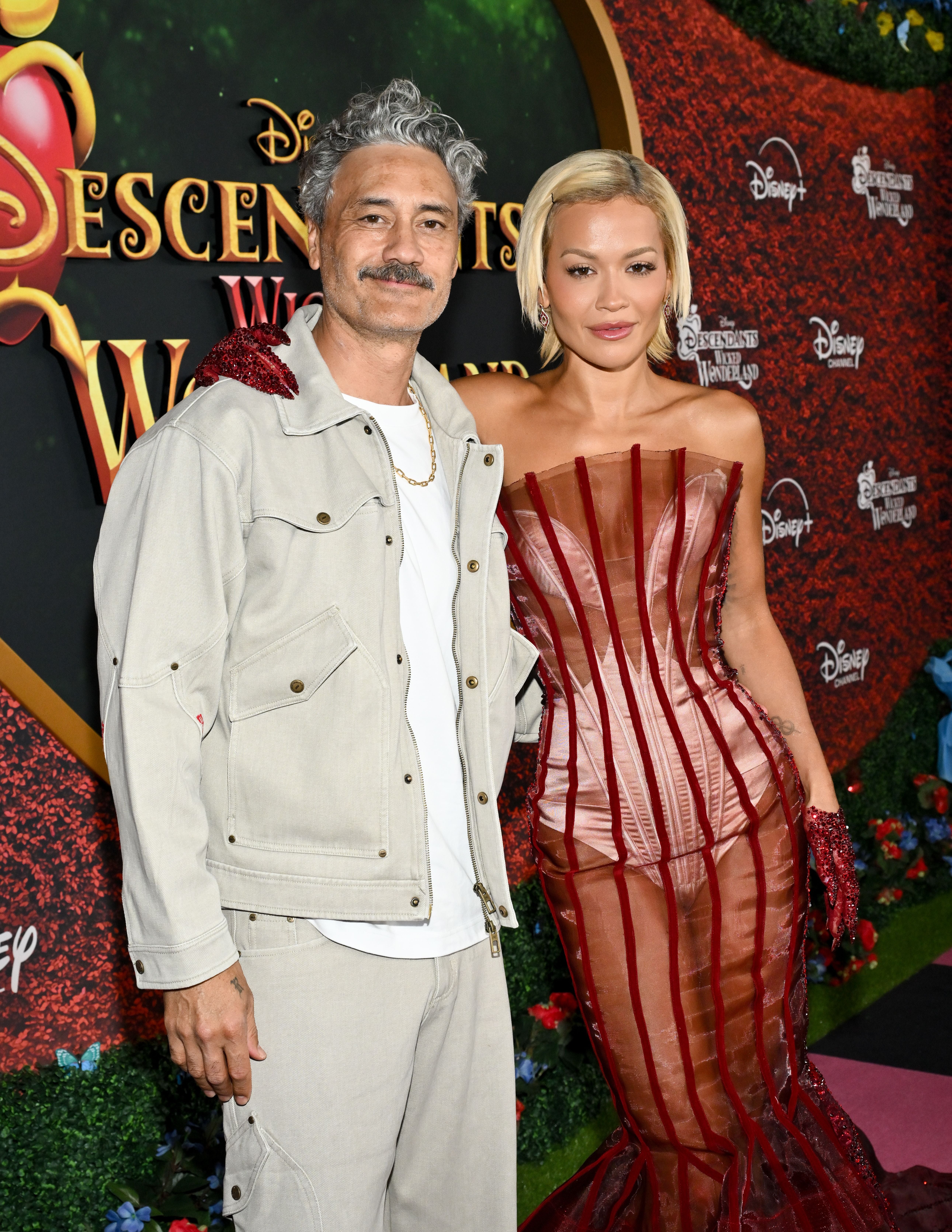 Ein Mann in hellen Kleidern und eine Frau im roten Kleid posieren auf einem Premieren-Teppich.