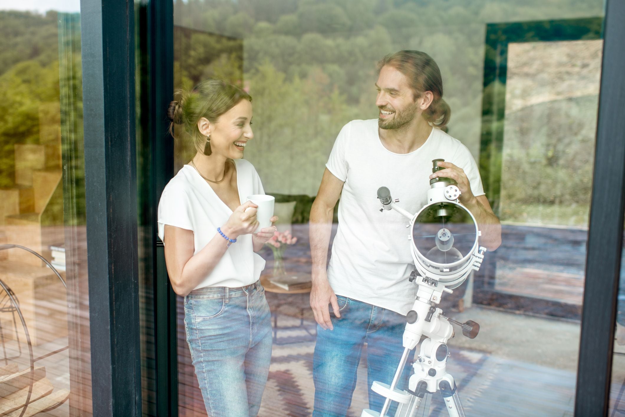 Junges Paar steht mit Teleskop am Fenster und lächelt sich an.
