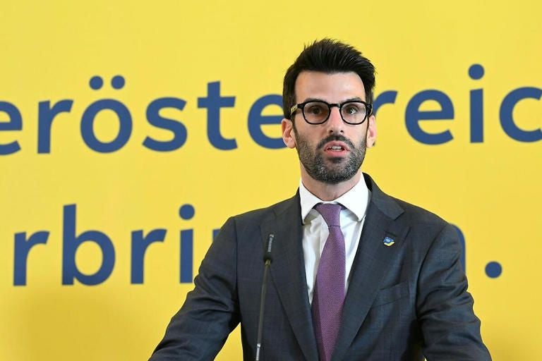Ein Mann im Anzug spricht vor einem gelben Hintergrund mit blauer Schrift bei einer Pressekonferenz.