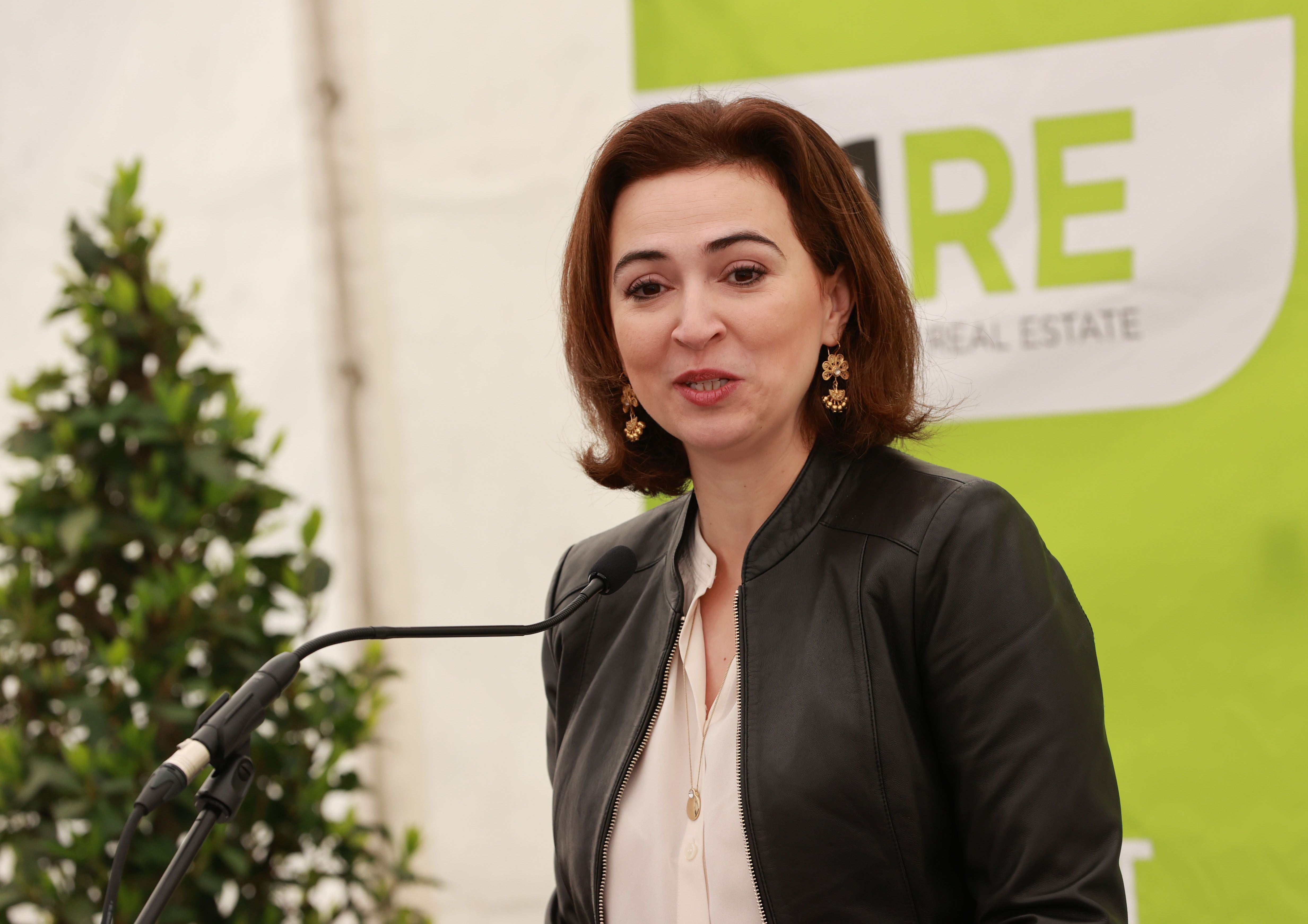 Eine Frau spricht bei einer Veranstaltung am Mikrofon, Hintergrund grün-weißes Banner.