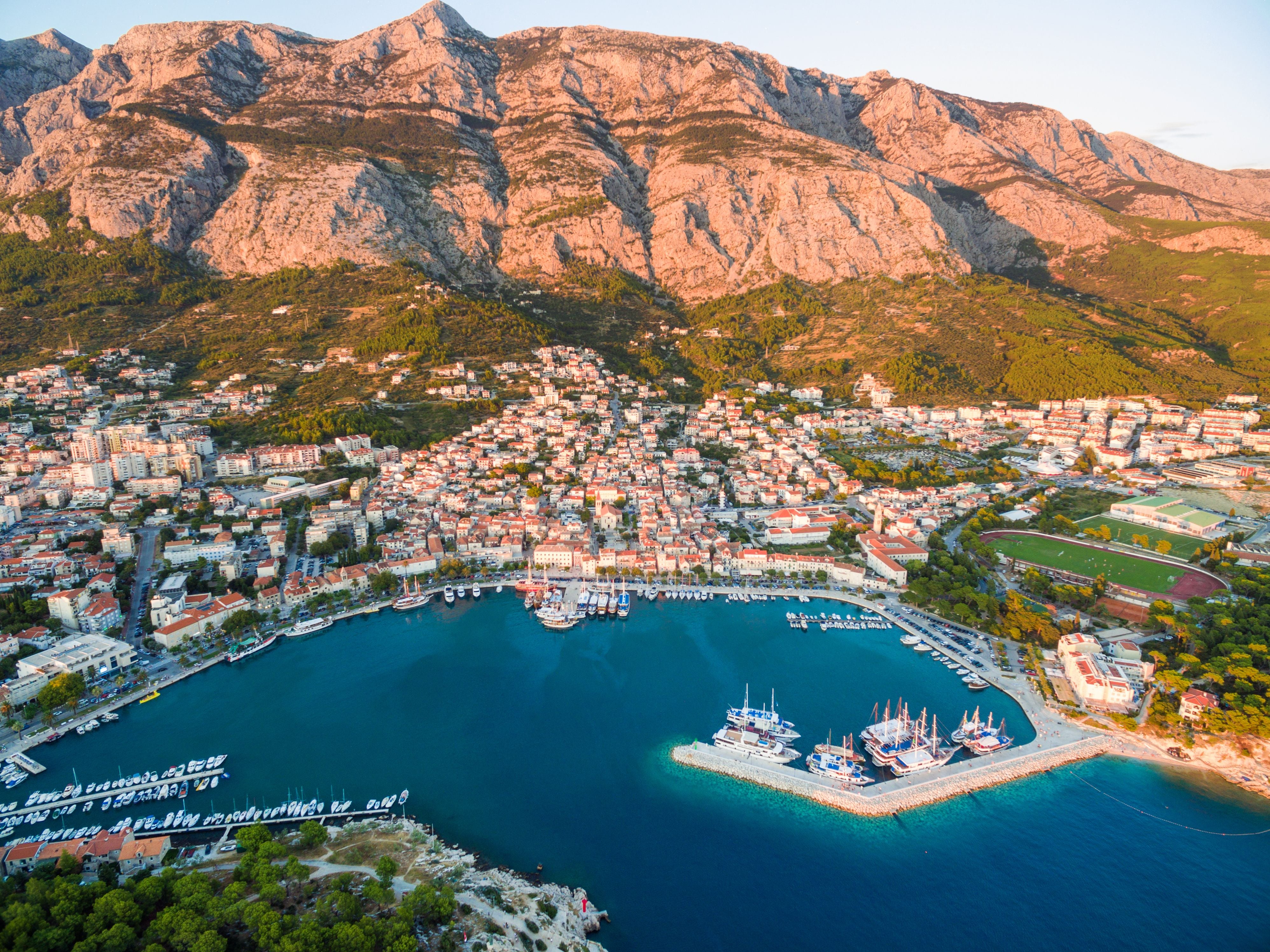 Luftaufnahme von Makarska mit Yachthafen und Biokovo-Gebirge im Hintergrund bei Sonnenuntergang.