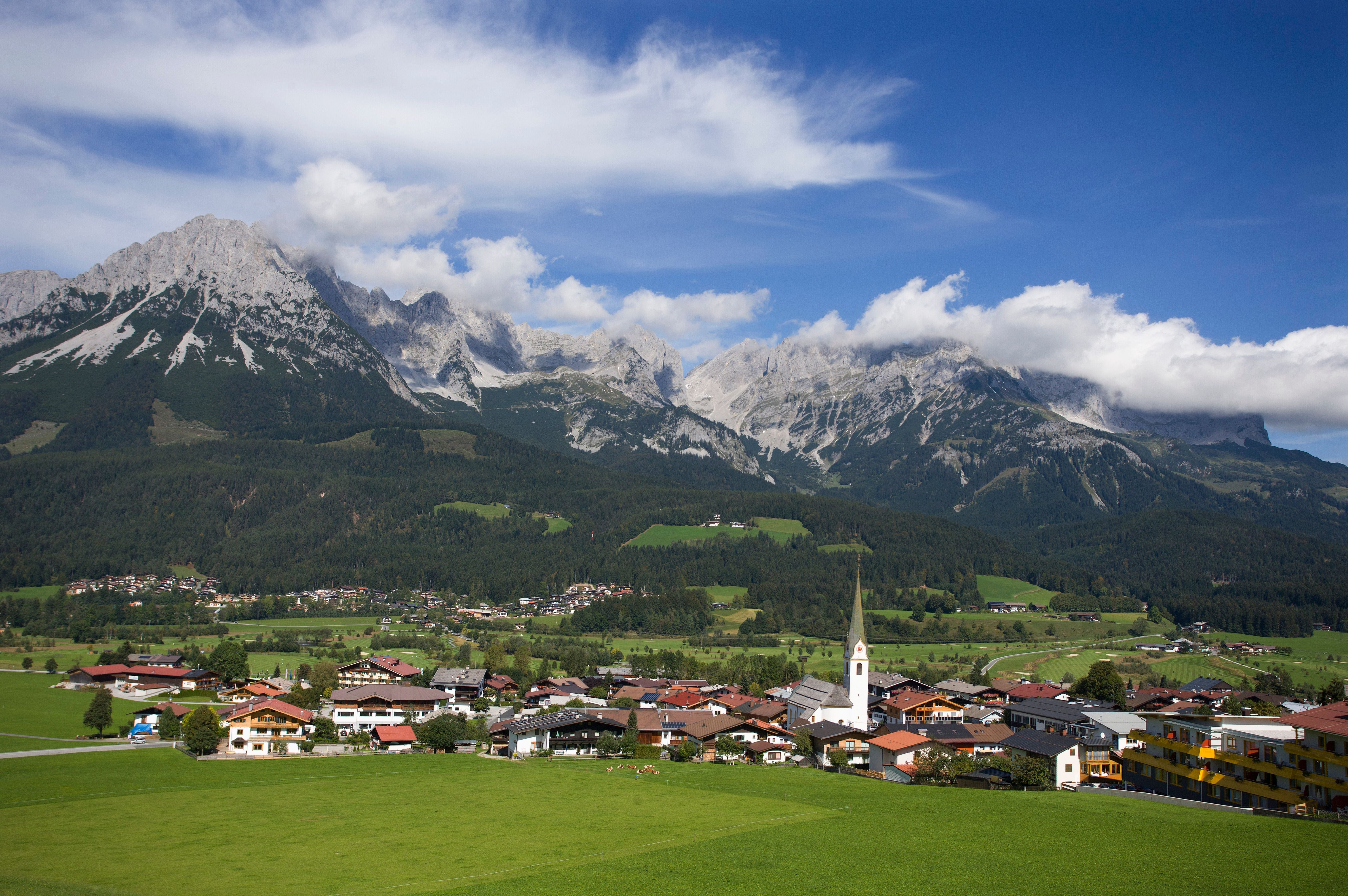 Blick auf das Dorf Ellmau mit Kirche vor den Alpen und grünen Wiesen unter blauem Himmel.