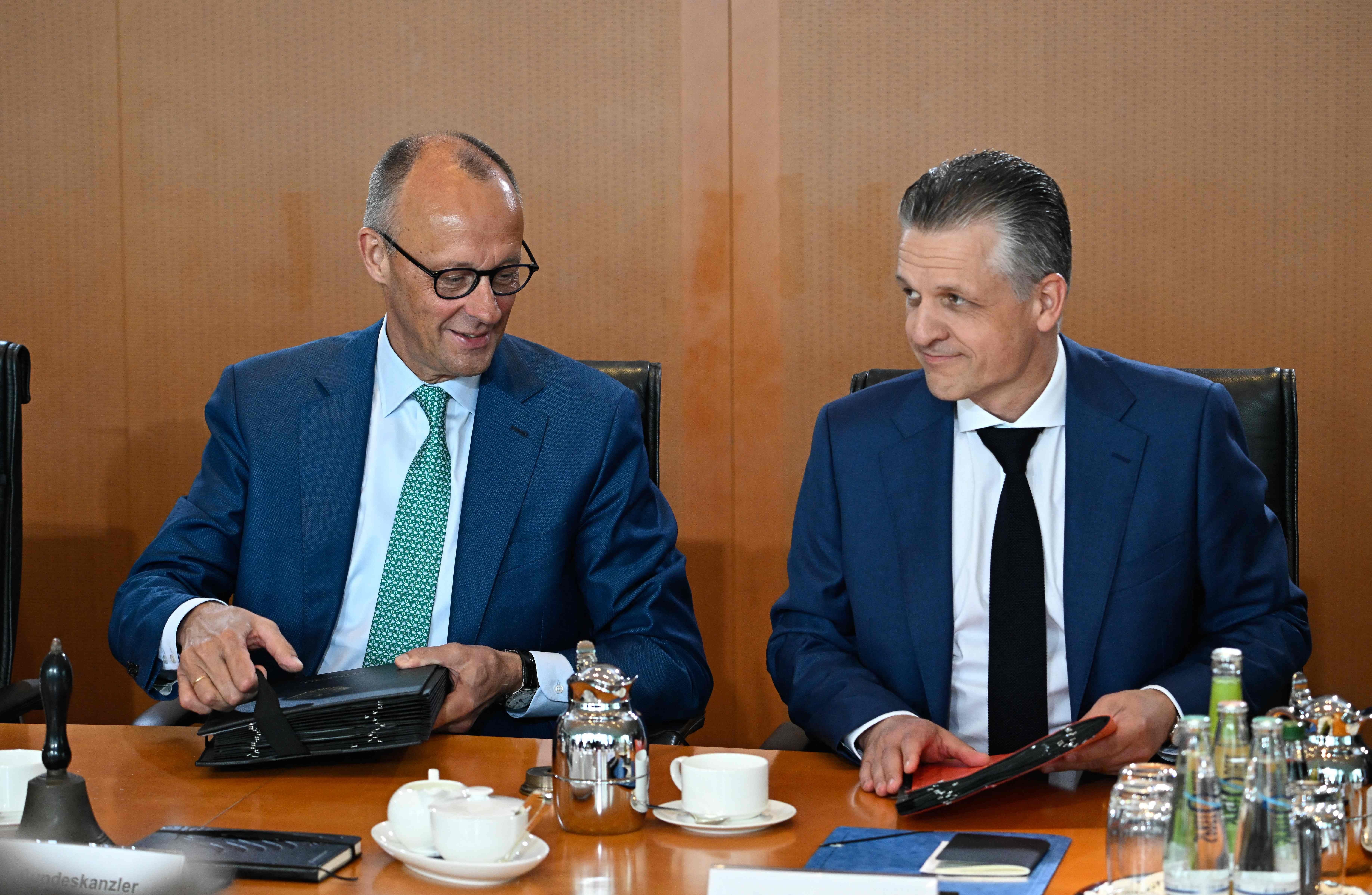 Zwei Männer im Anzug sitzen an einem Konferenztisch mit Unterlagen und Getränken.