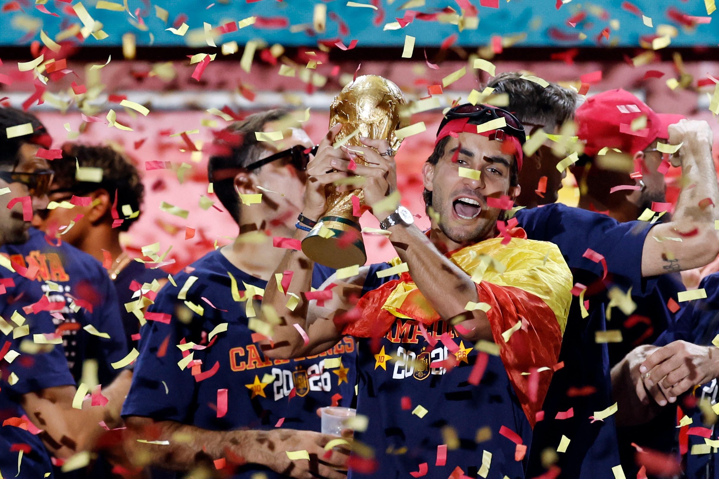 Spanischer Spieler feiert mit WM-Pokal, umgeben von Konfetti und jubelnden Teamkollegen.