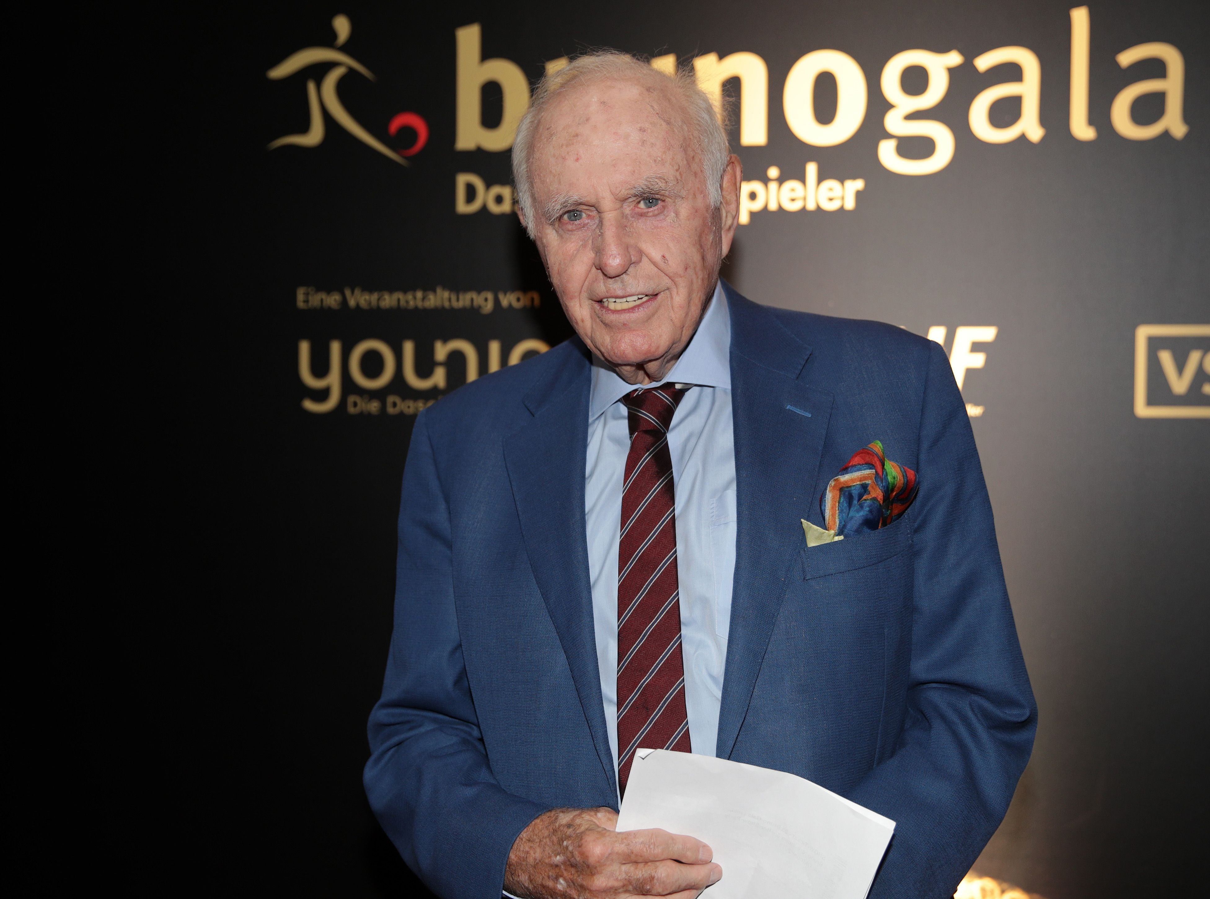Ein älterer Mann im blauen Anzug steht bei einer Gala vor einer Wand mit goldener Schrift.