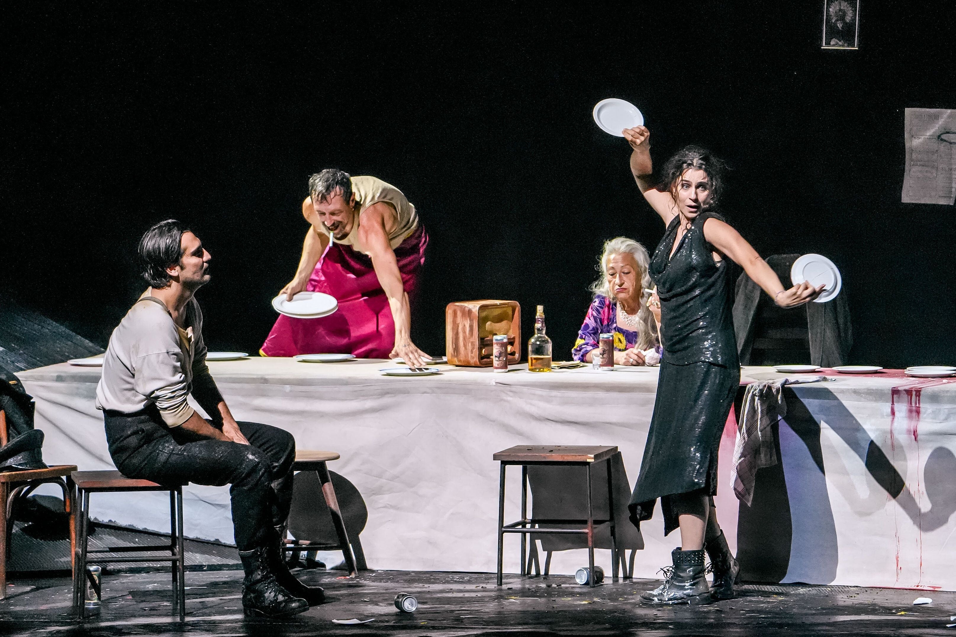 Vier Schauspieler:innen inszenieren eine theatralische Szene an einem gedeckten Tisch mit Tellern und Getränken.