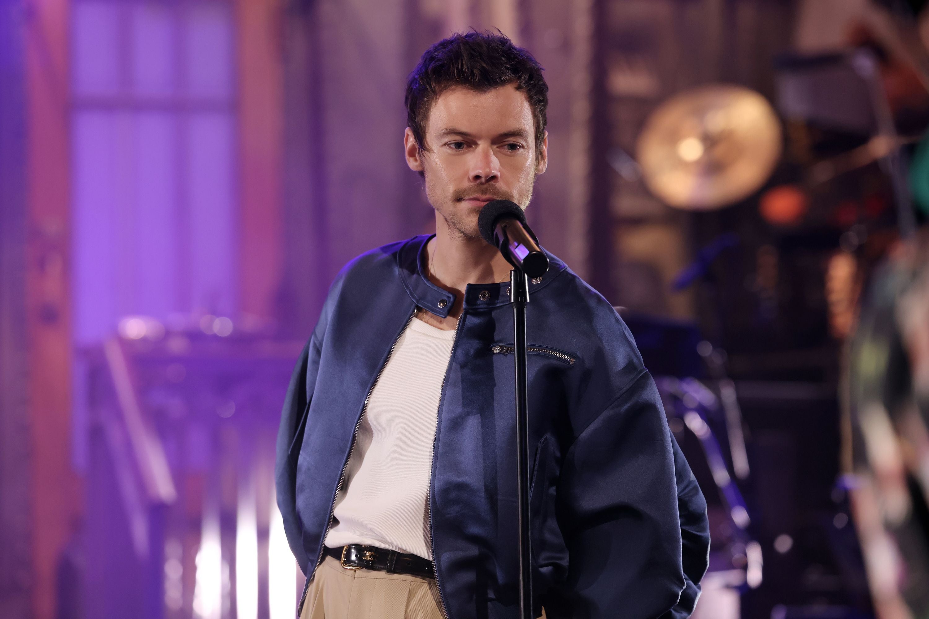 Ein Mann singt auf einer Bühne in lila Licht und trägt eine blaue Jacke und ein weißes Shirt.