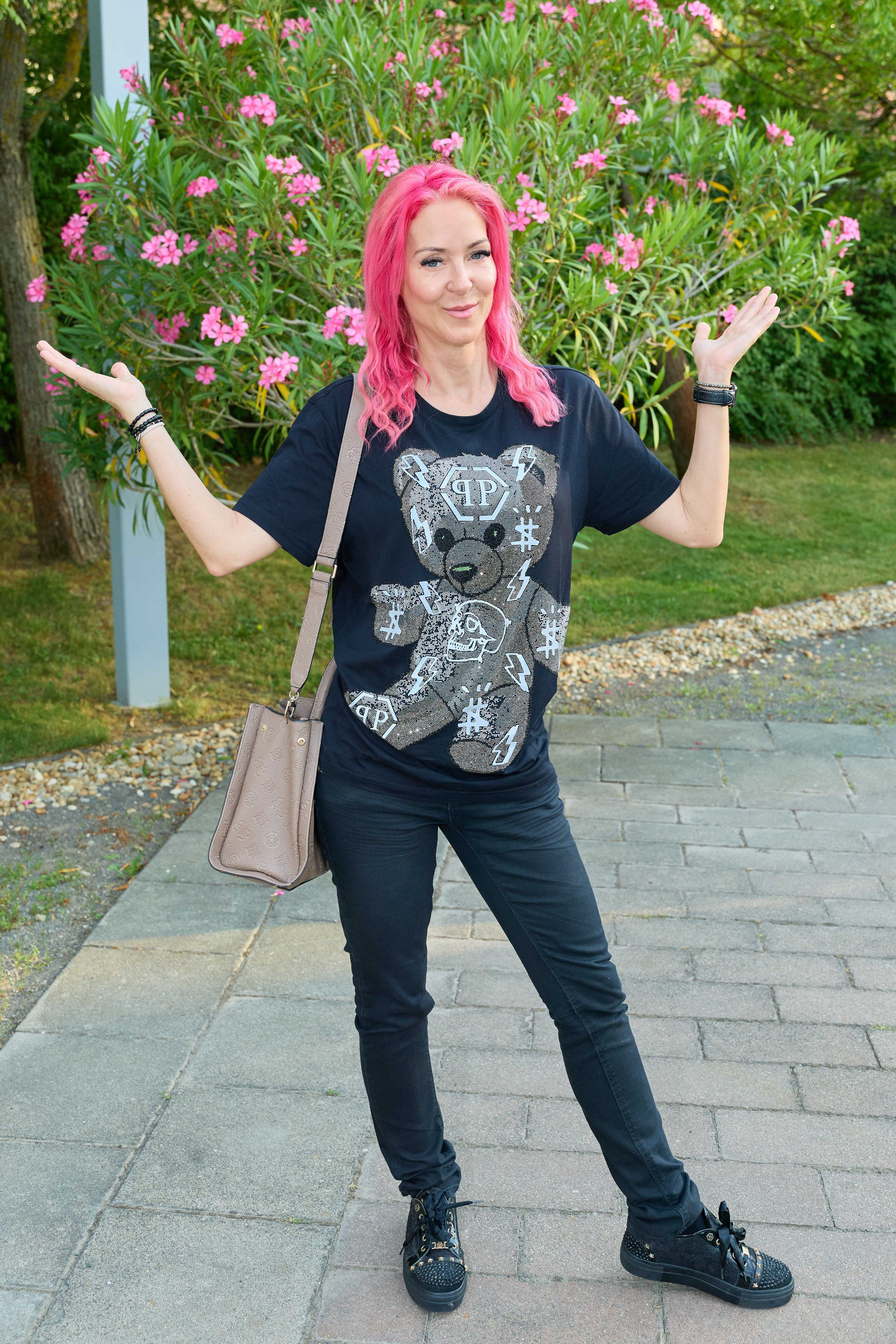Frau mit pinken Haaren, schwarzem Teddybär-Shirt und Handtasche posiert draußen vor Blumen.