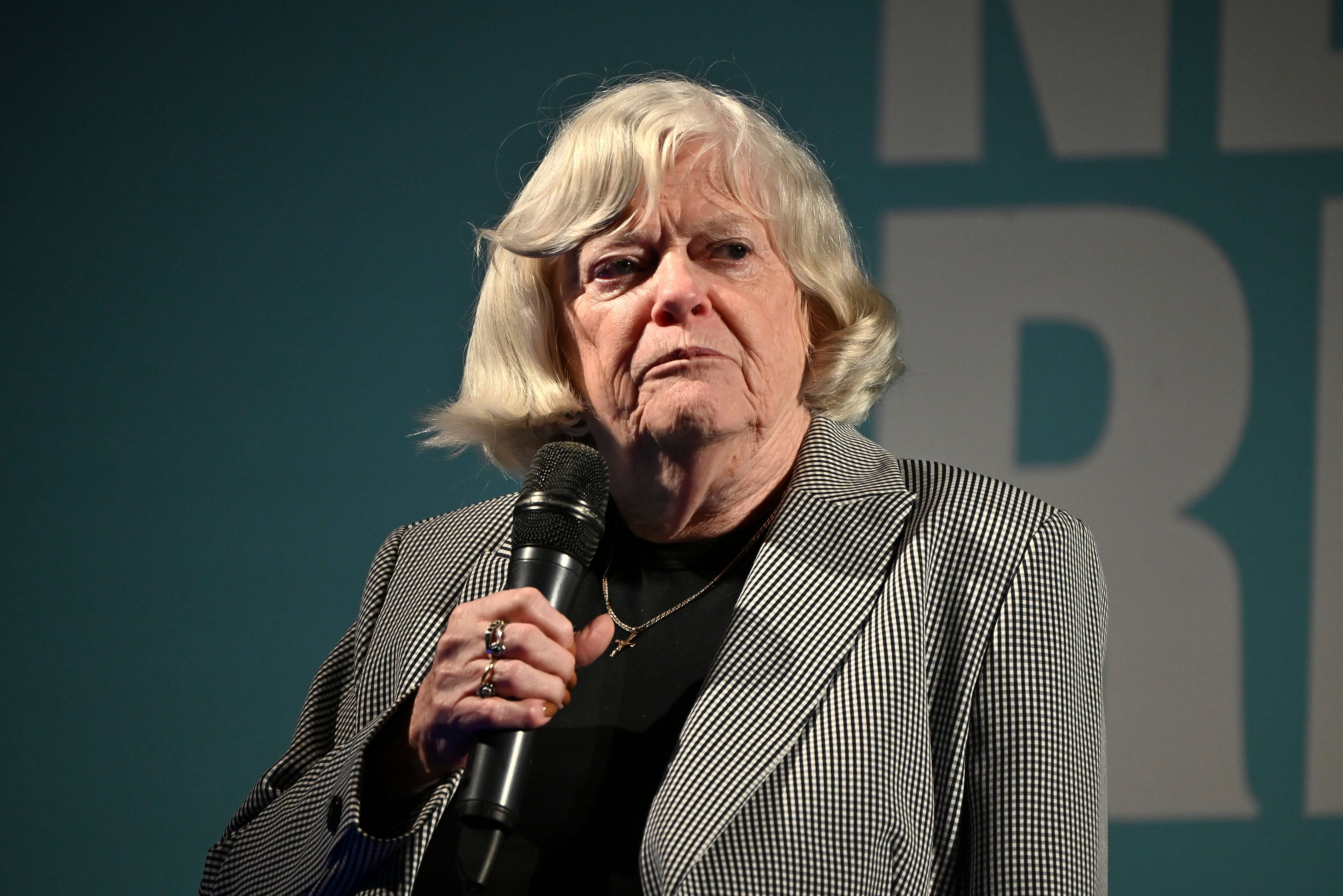 Ältere Frau mit blondem Haar spricht bei einer Konferenz und hält ein Mikrofon in der Hand.