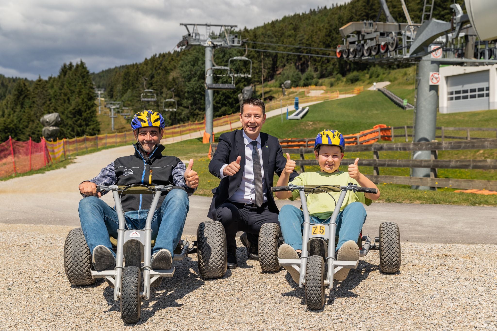 Zwei Personen auf Mountaincarts und ein Mann in Anzug zeigen Daumen hoch im Freizeitpark.