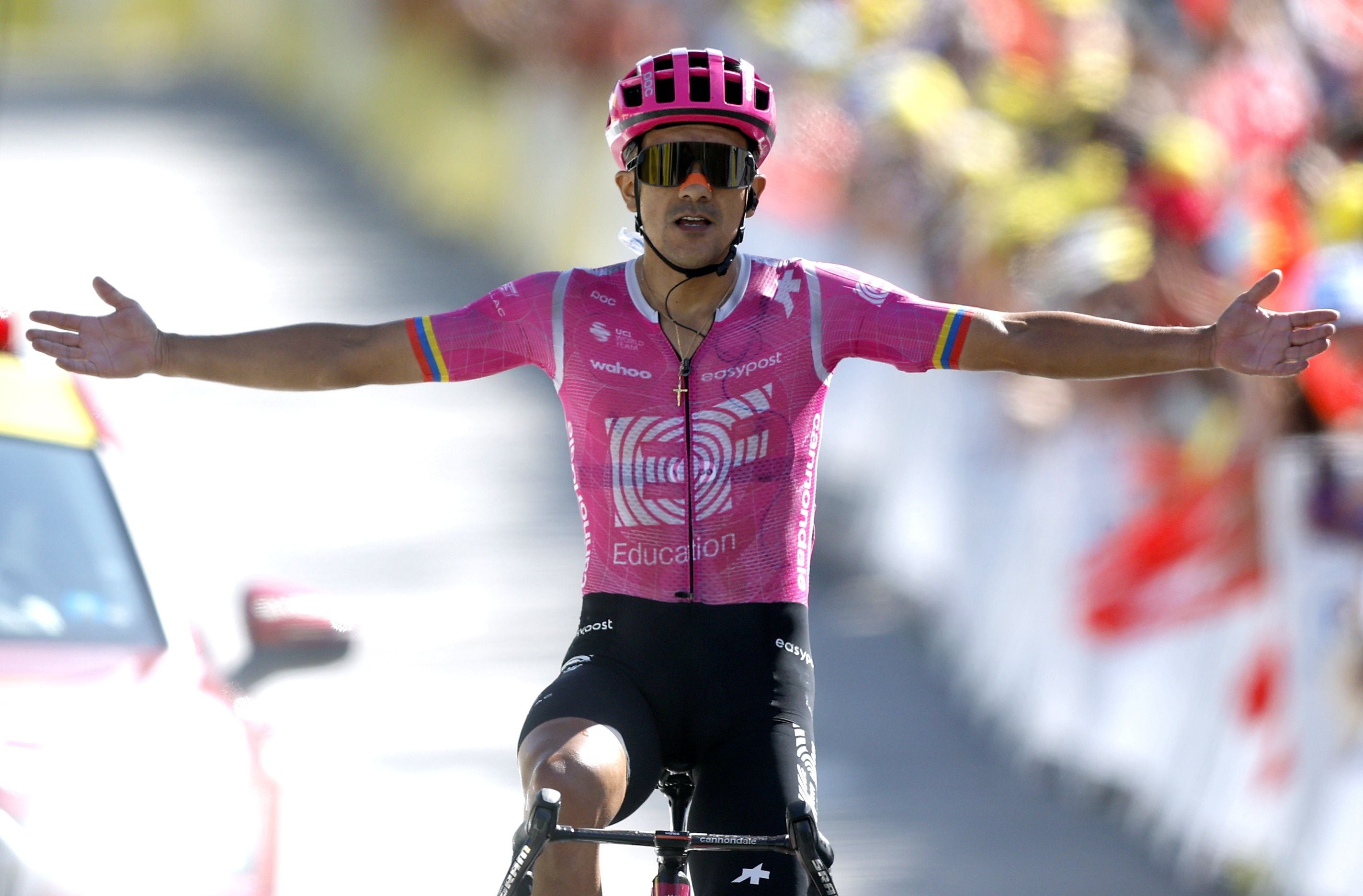 Ein Radprofi im rosa Trikot fährt auf seinem Rennrad mit ausgebreiteten Armen und geöffnetem Mund über die Ziellinie einer Straße. Im Hintergrund sind Zuschauer und die Straße zu sehen.