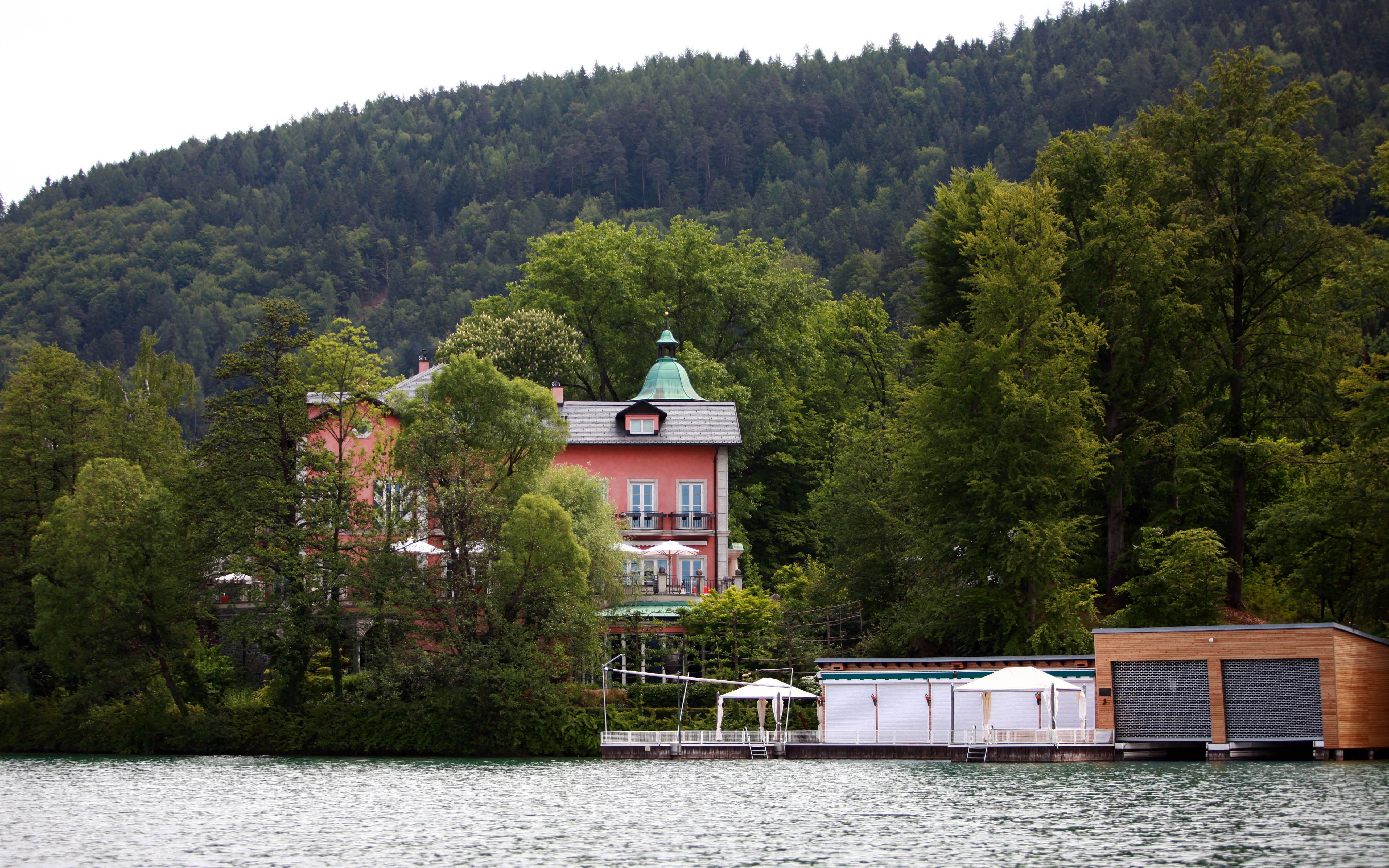 Villa am Seeufer, von Bäumen umgeben, mit Bootshaus und Pavillon am Wasser, bewaldeter Hügel im Hintergrund.