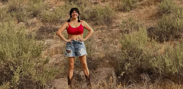 Frau in rotem Top und Jeansshorts steht lächelnd in trockener, buschiger Landschaft.