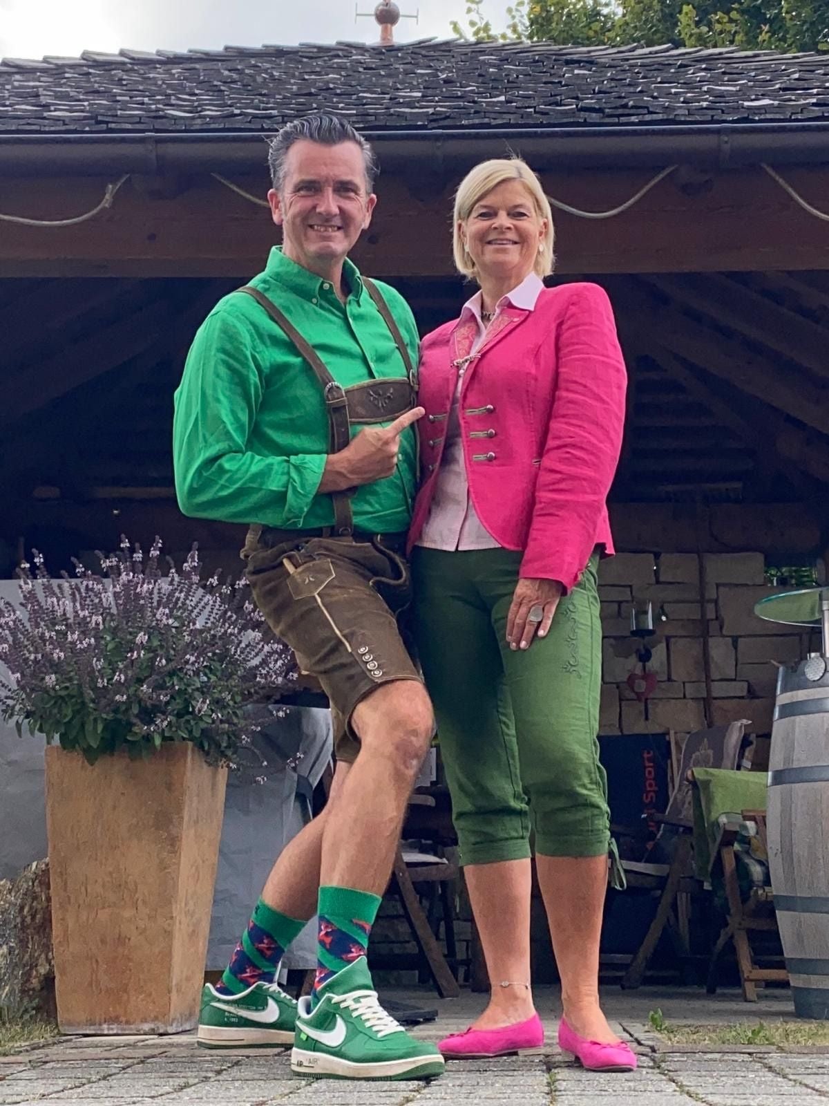 Ein Mann in Lederhose und eine Frau in Trachtenjacke posieren lächelnd im Freien.
