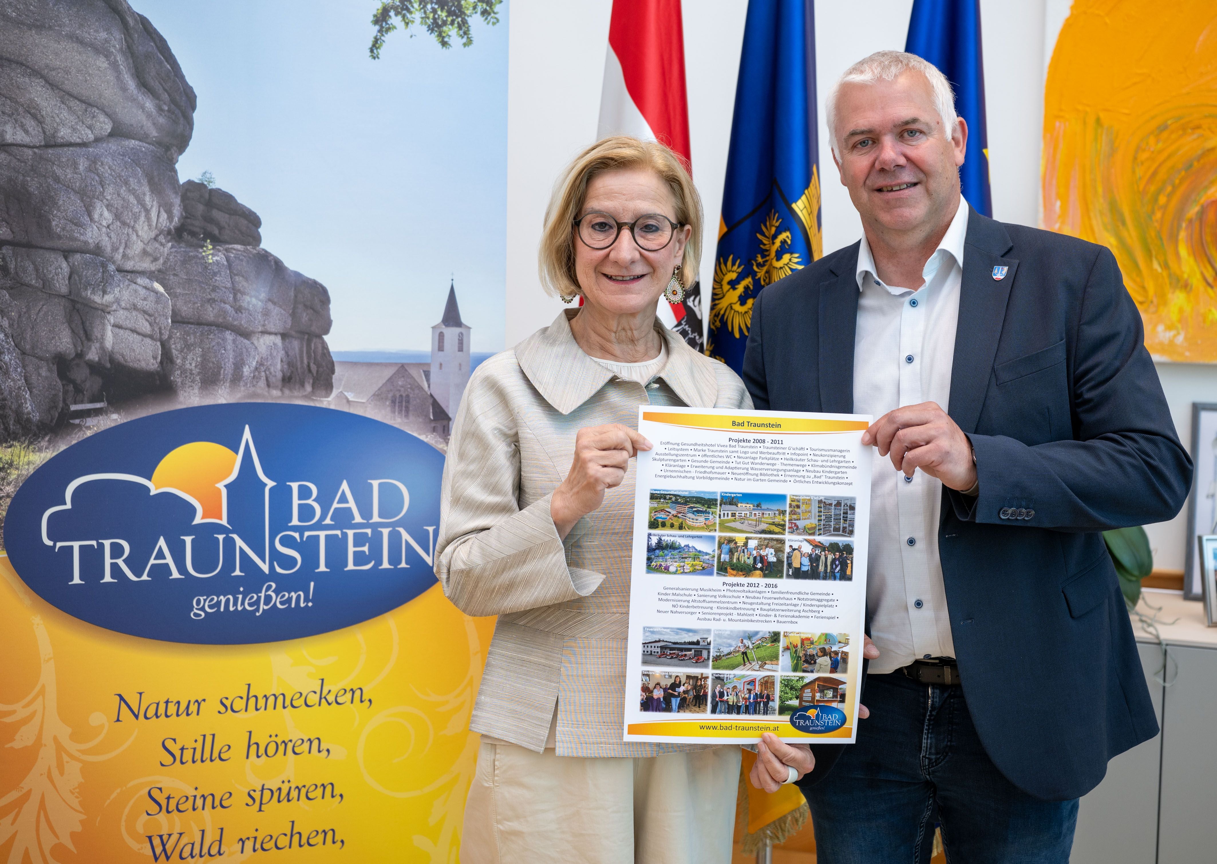 Zwei Personen halten ein Informationsblatt über Bad Traunstein vor Werbebanner und Fahnen.