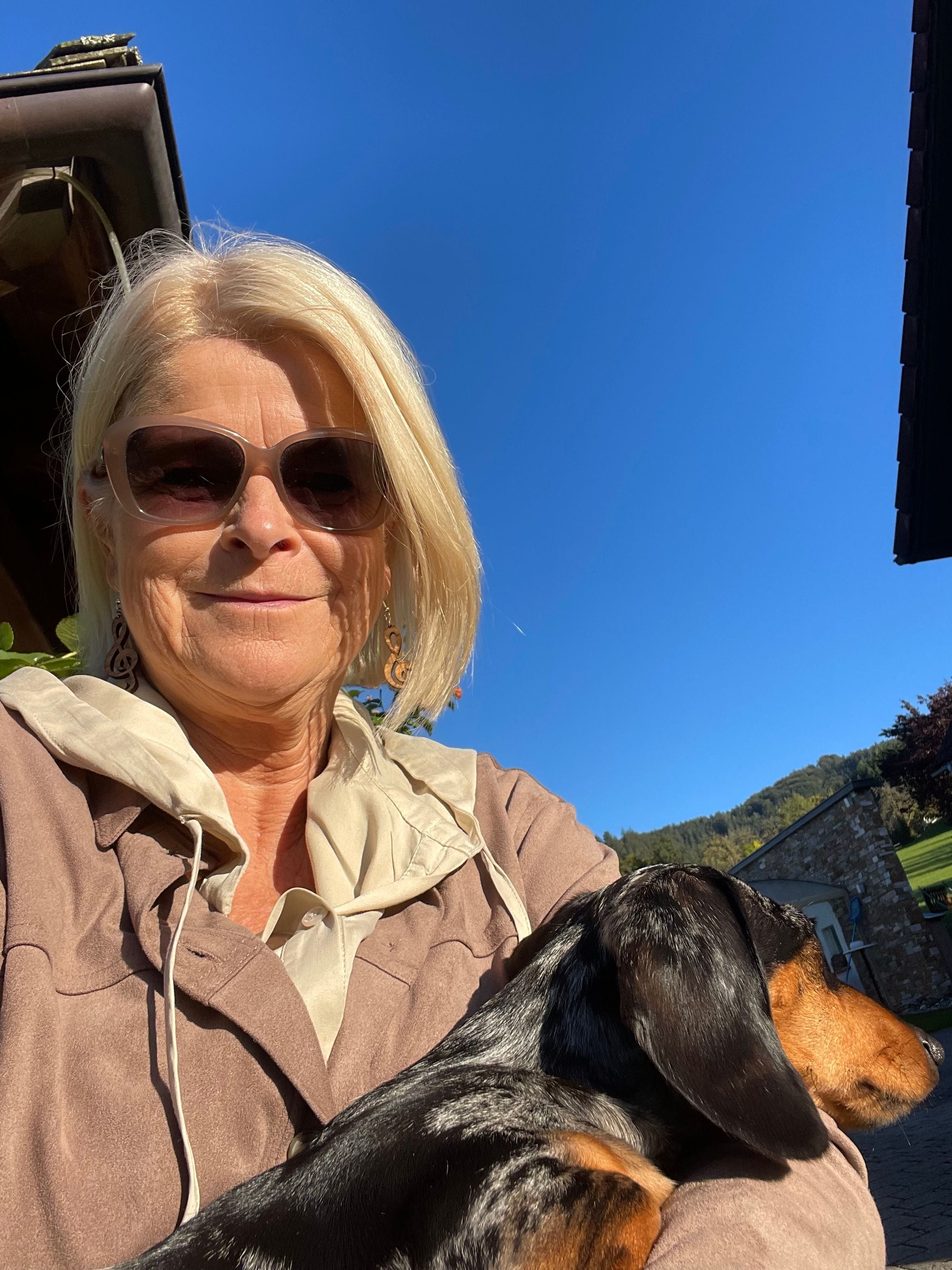 Frau mit Sonnenbrille hält einen Dackel im Arm, blauer Himmel und Berge im Hintergrund.