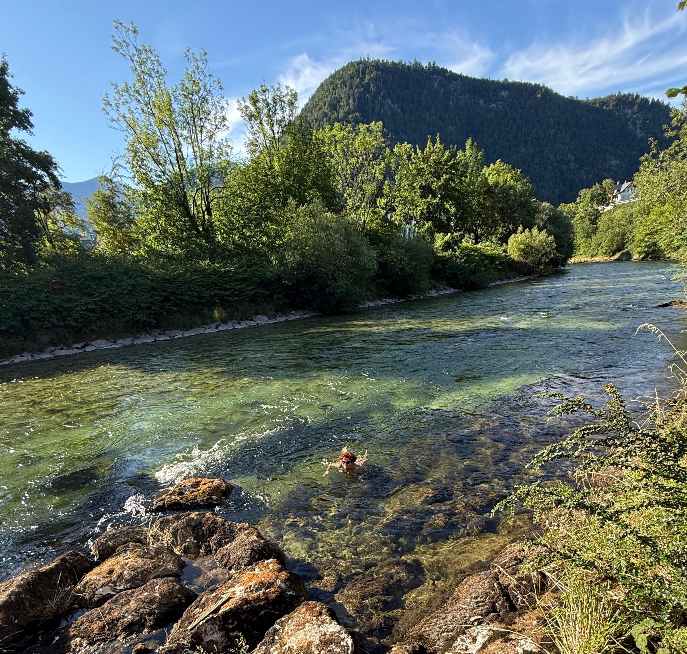 Eine Person schwimmt in einem klaren Fluss, umgeben von Bäumen und Bergen im Hintergrund.
