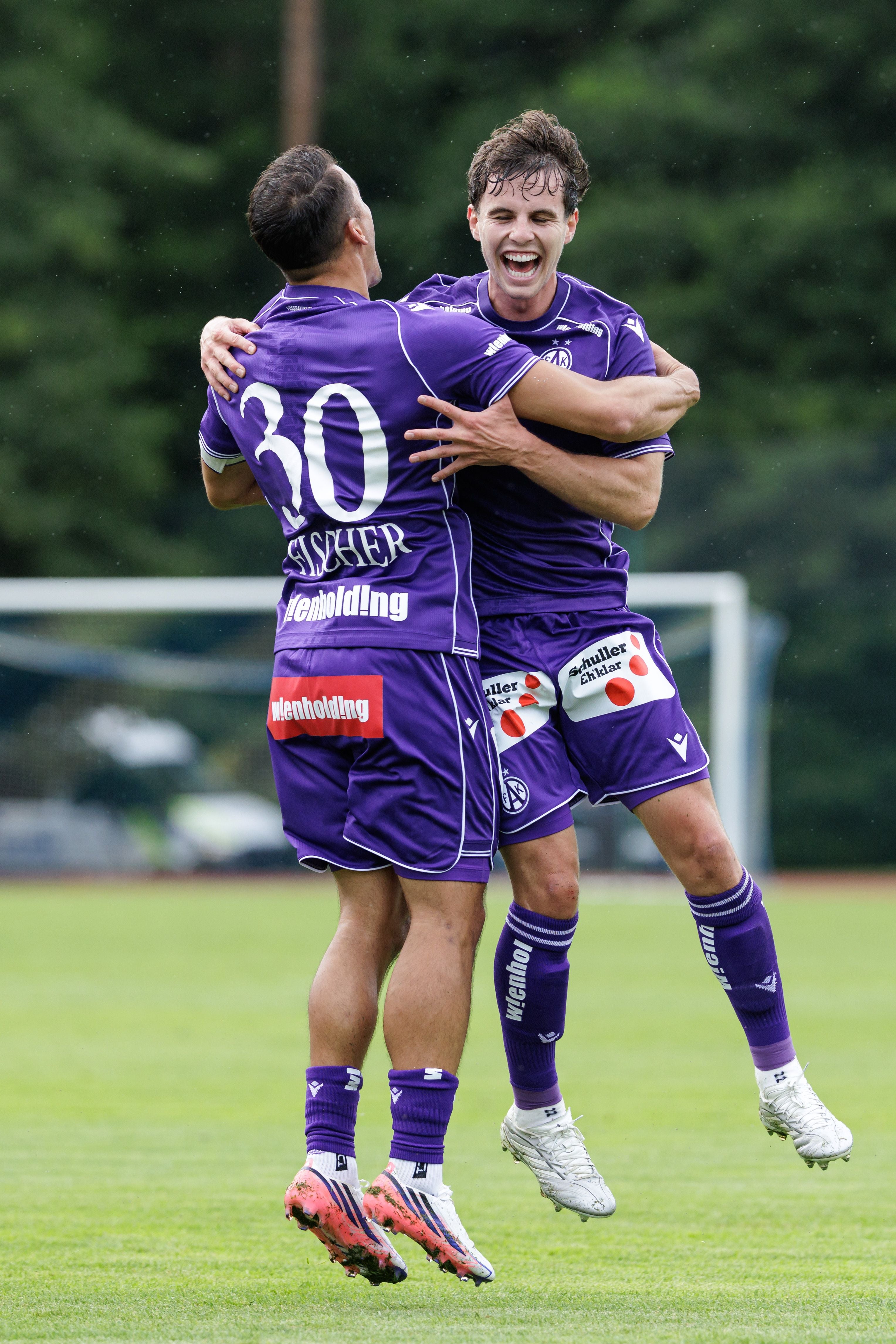 Zwei Fußballspieler von Austria Wien im violetten Trikot springen auf dem Rasen in die Luft und umarmen sich freudig. Der linke Spieler ist von hinten zu sehen, auf seinem Trikot steht die Nummer 30 und der Name Fischer.