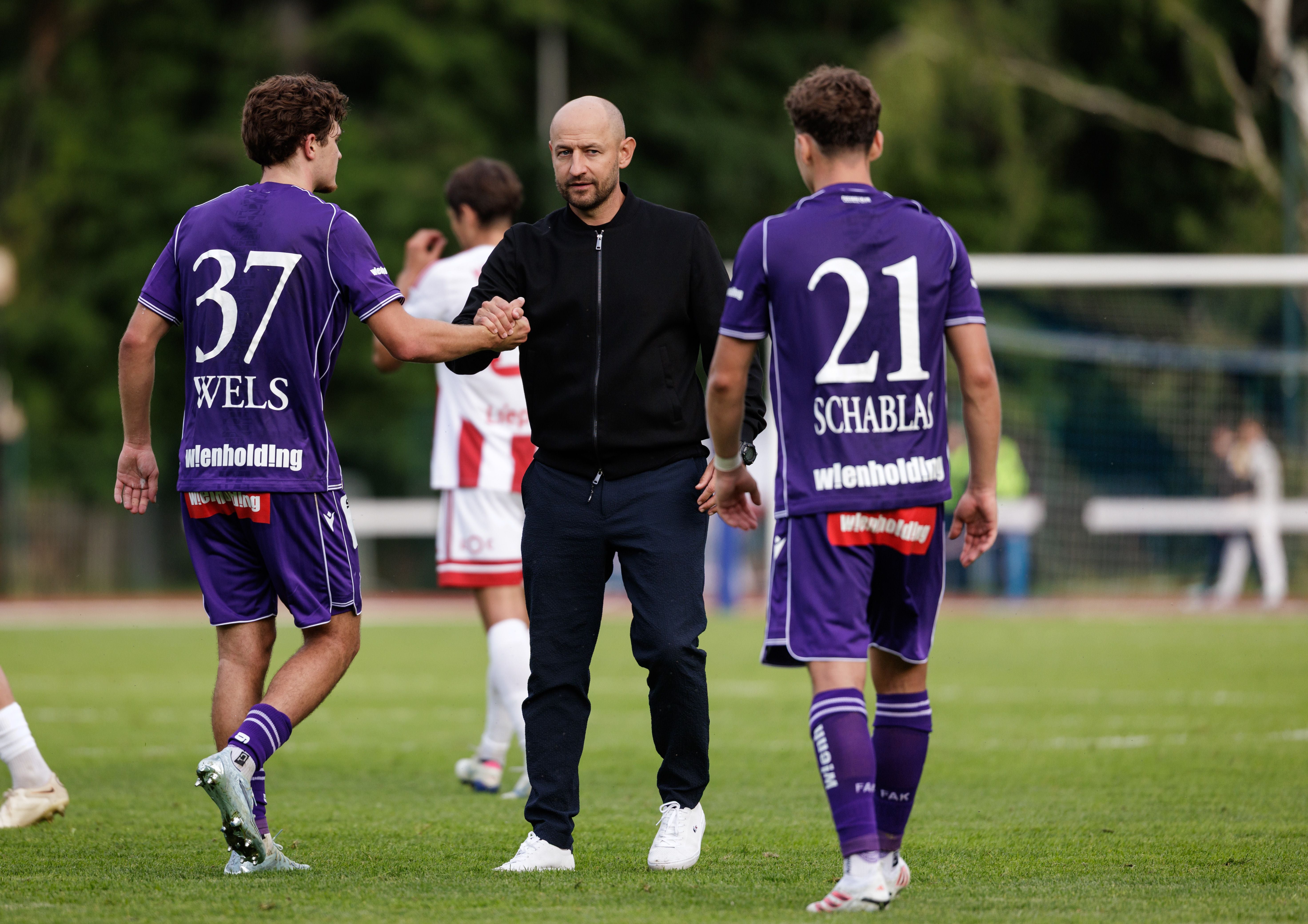 Ein glatzköpfiger Trainer in dunkler Kleidung steht in der Mitte eines Fußballfeldes und gibt einem Spieler im violetten Trikot mit der Rückennummer 37 den Handschlag. Ein weiterer Spieler im gleichen Trikot mit der Nummer 21 steht daneben.