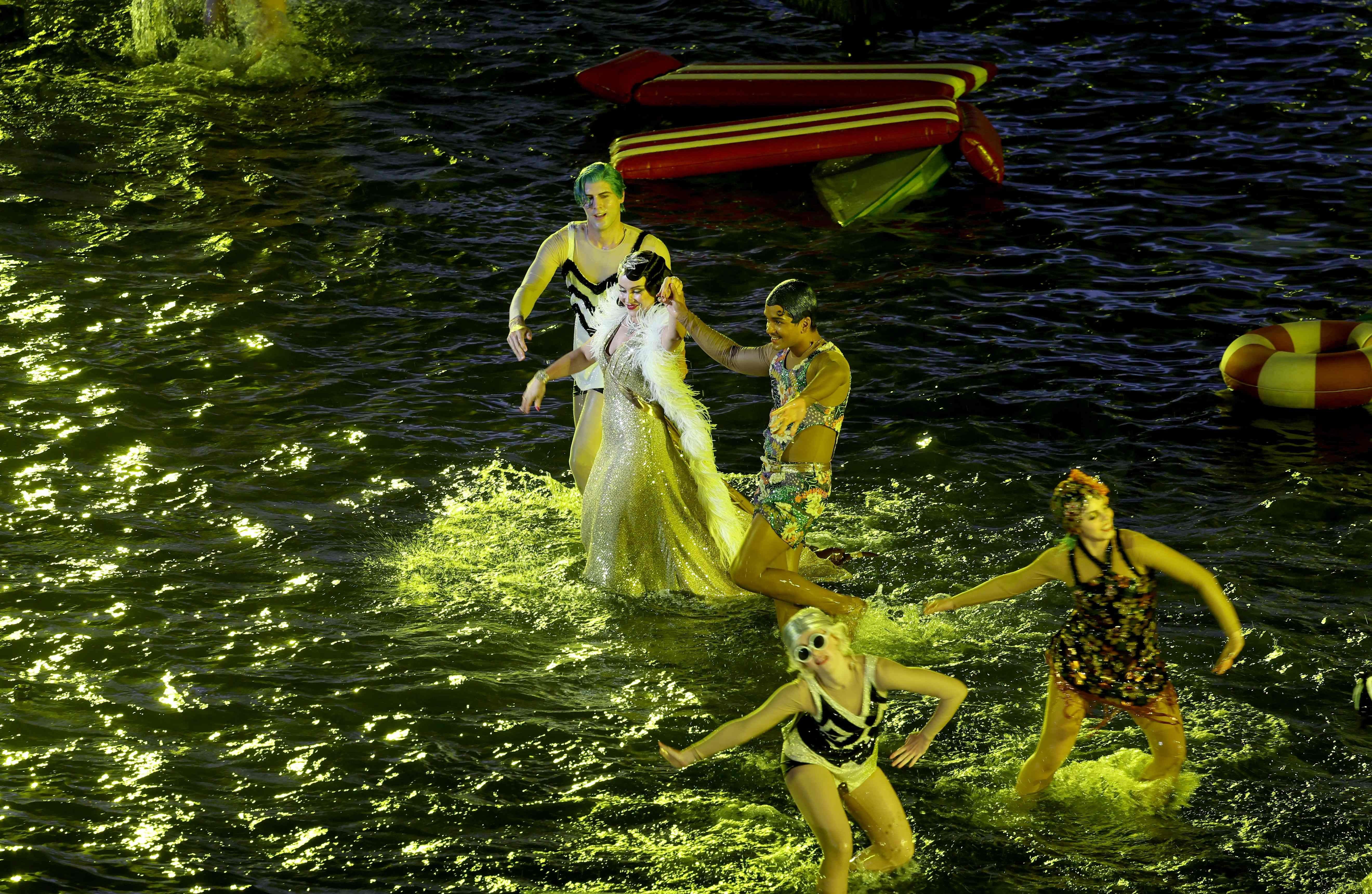 Mehrere Darsteller tanzen in bunten Kostümen bei Nacht im Wasser auf einer beleuchteten Bühne.