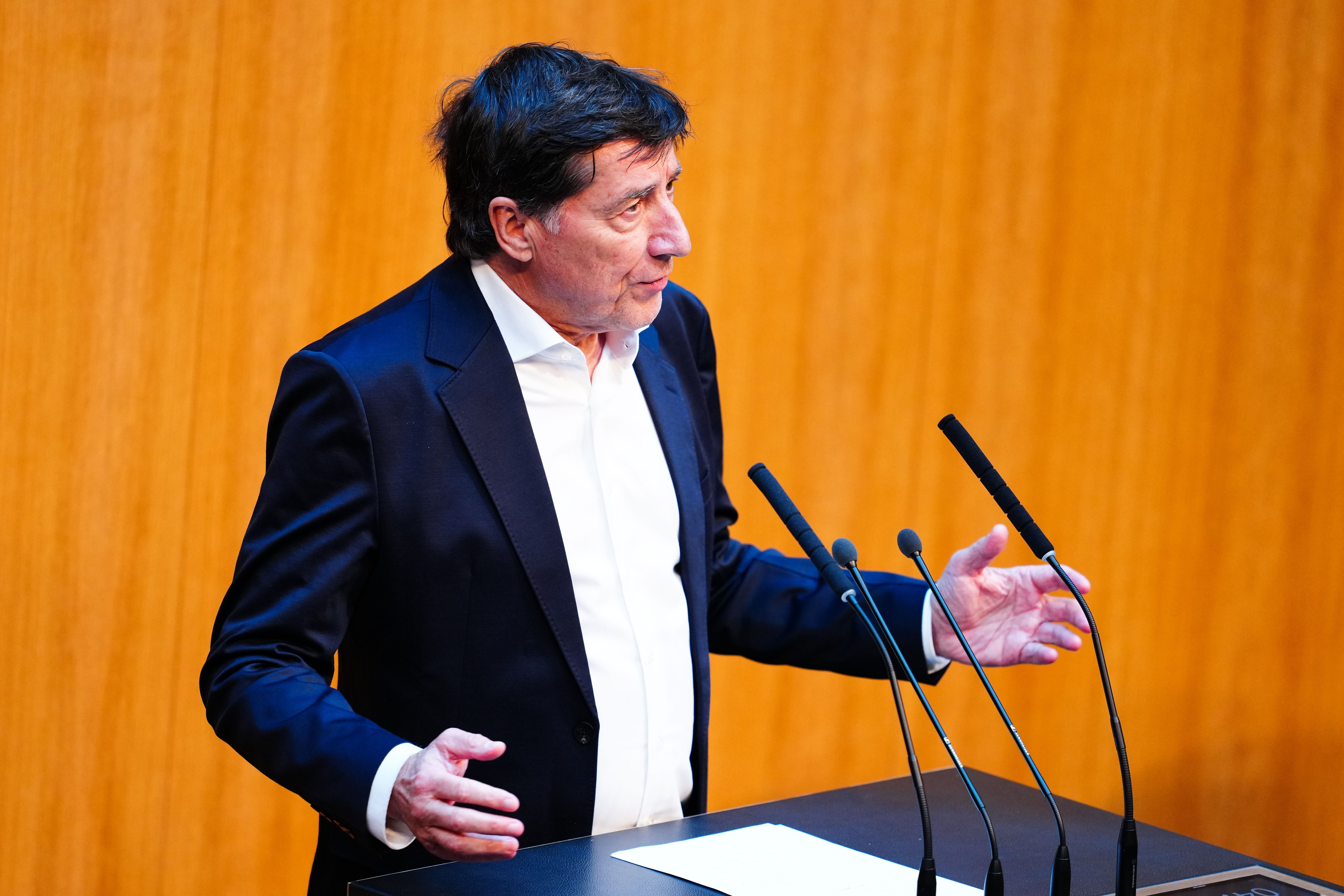Mann im Anzug spricht an einem Rednerpult mit Mikrofonen vor Holzvertäfelung.