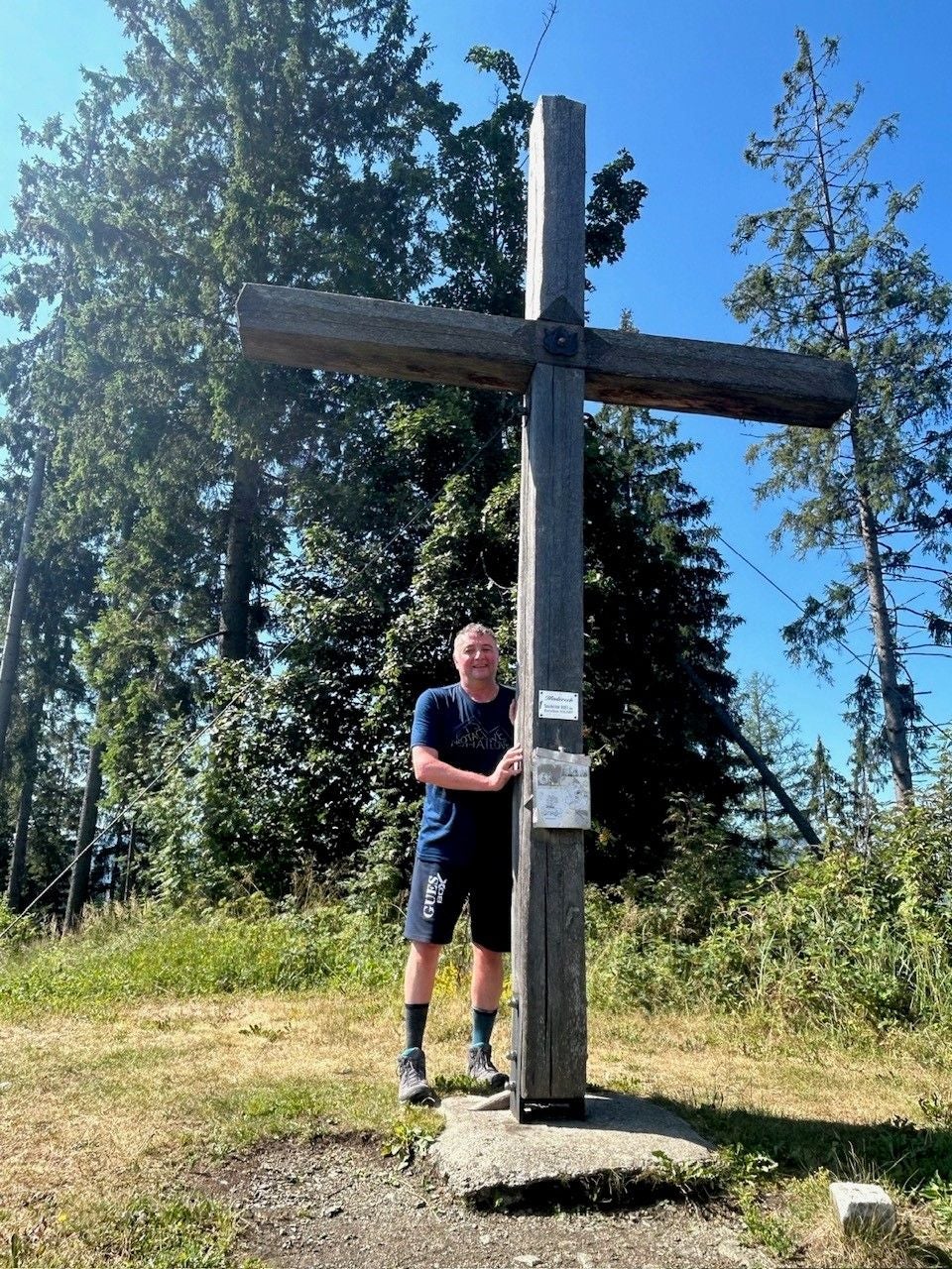 Ein Mann steht bei Sonnenschein an einem hölzernen Gipfelkreuz im Wald.
