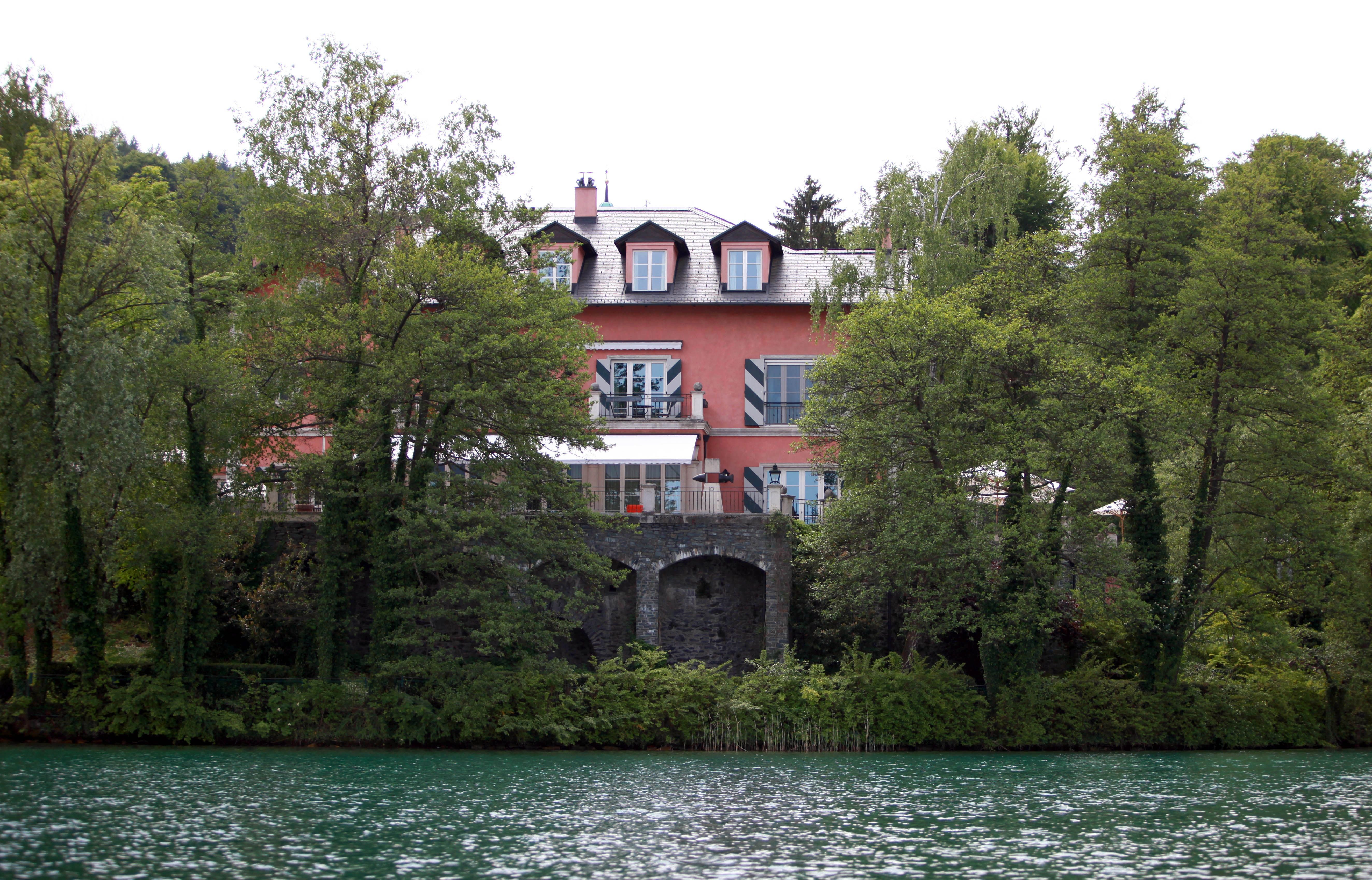 Große Villa mit rosa Fassade hinter Bäumen am Ufer eines Sees.