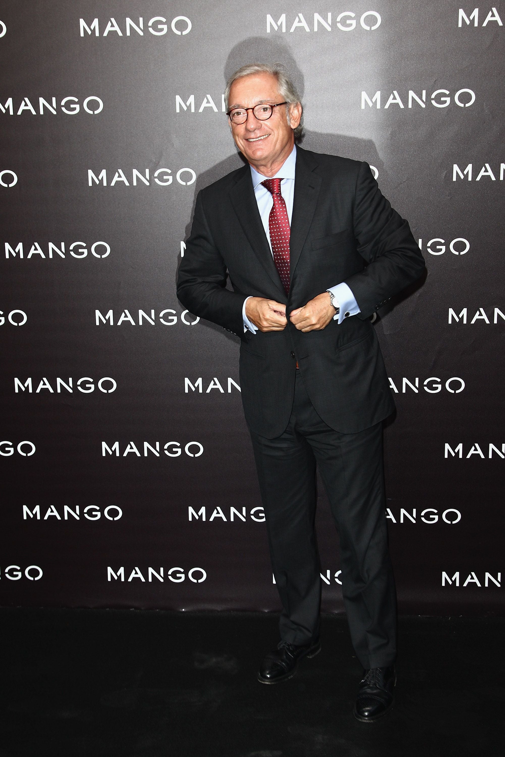 Ein älterer Mann im Anzug steht vor einer Mango-Fotowand und lächelt in die Kamera.