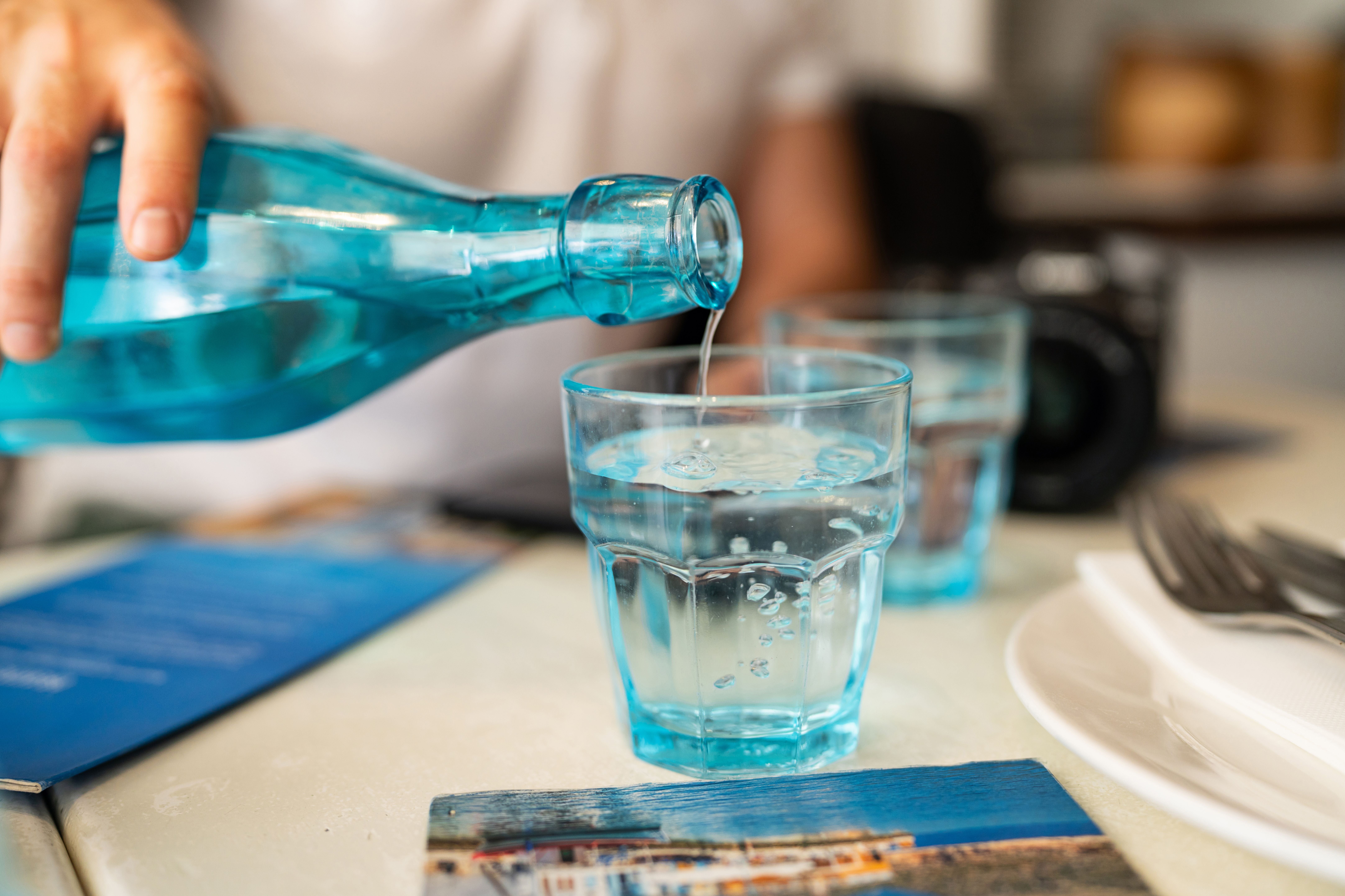 Wasser wird aus einer blauen Flasche in ein Trinkglas auf einem gedeckten Tisch gegossen.