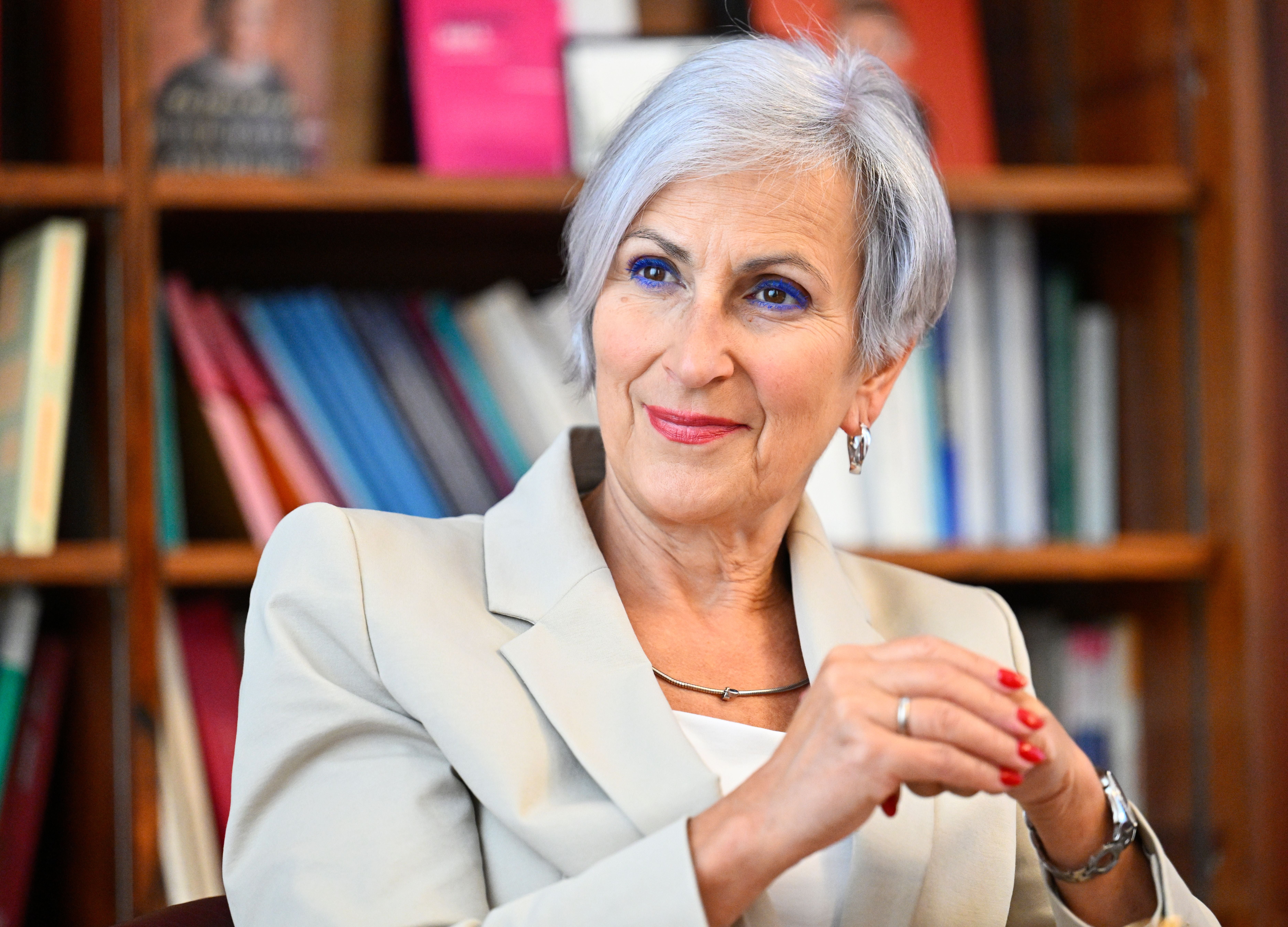 Eine ältere Frau mit grauen Haaren und hellem Blazer sitzt vor einem Bücherregal und lächelt.