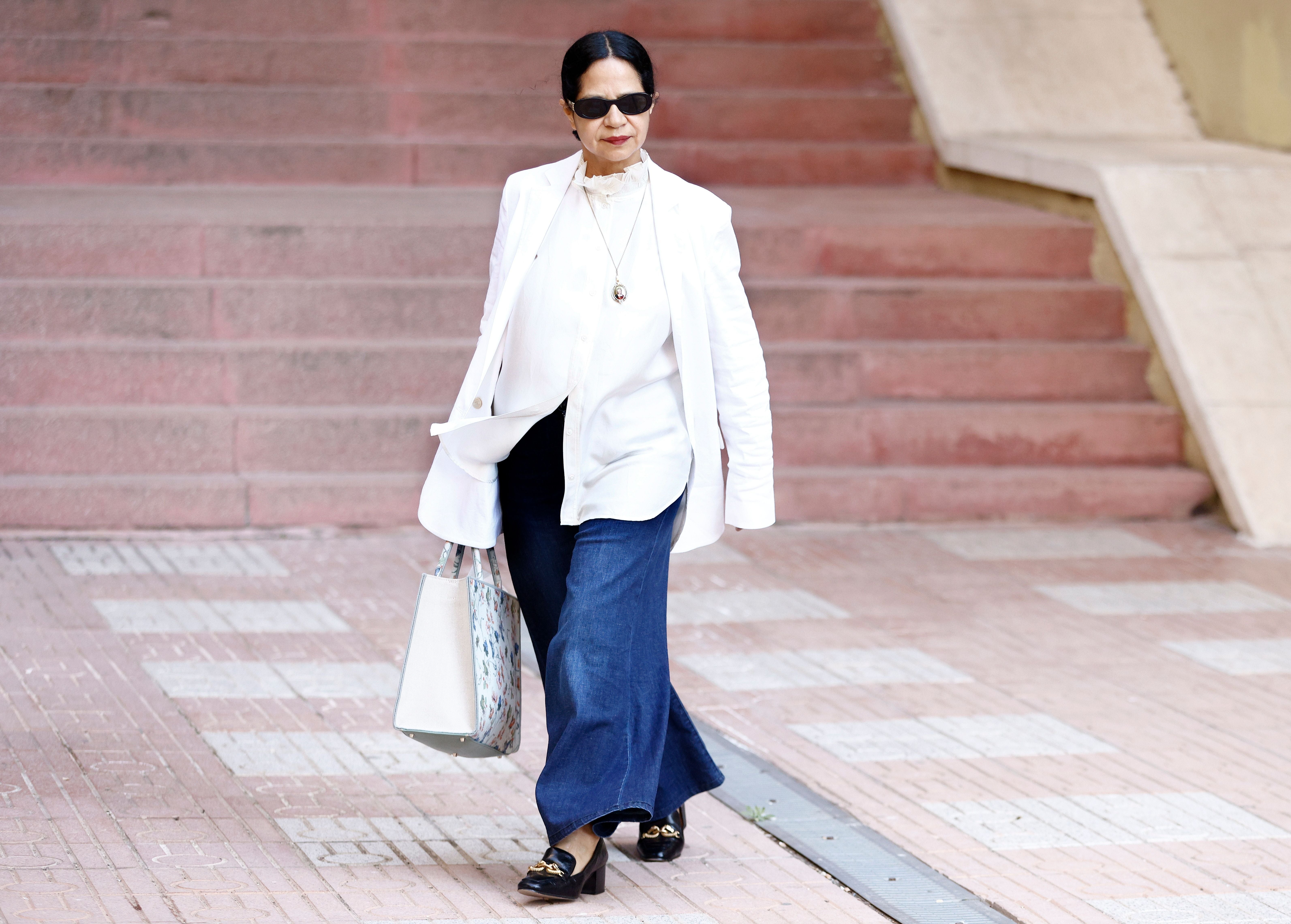 Eine Frau in weißem Blazer, blauer Hose und Sonnenbrille läuft auf einem Gehweg.