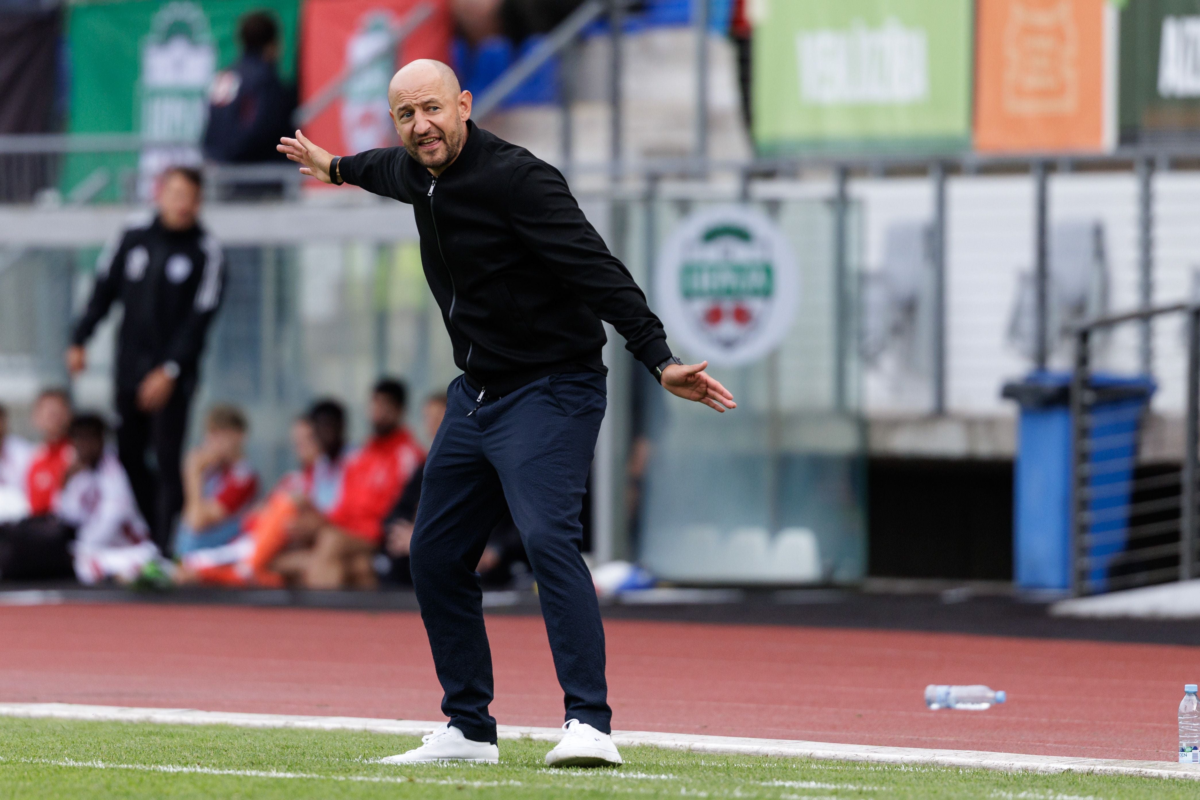 Ein glatzköpfiger Mann im schwarzen Jackett, dunkler Hose und weißen Turnschuhen steht am Rand eines Fußballfeldes. Er beugt sich leicht nach vorne und gibt mit ausgebreiteten Armen Anweisungen.