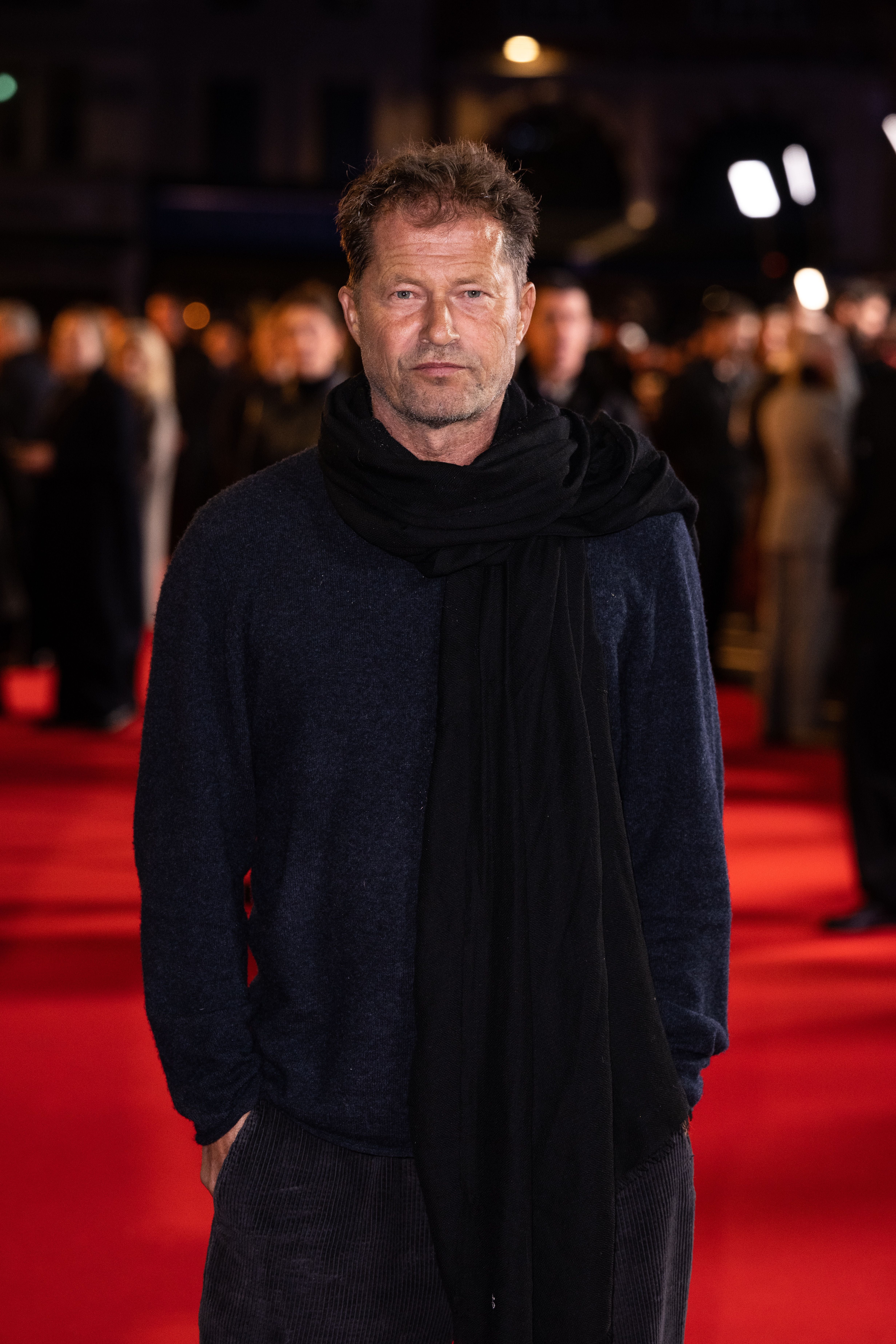 Ein Mann mit dunkelblauem Pullover und schwarzem Schal steht auf einem roten Teppich.