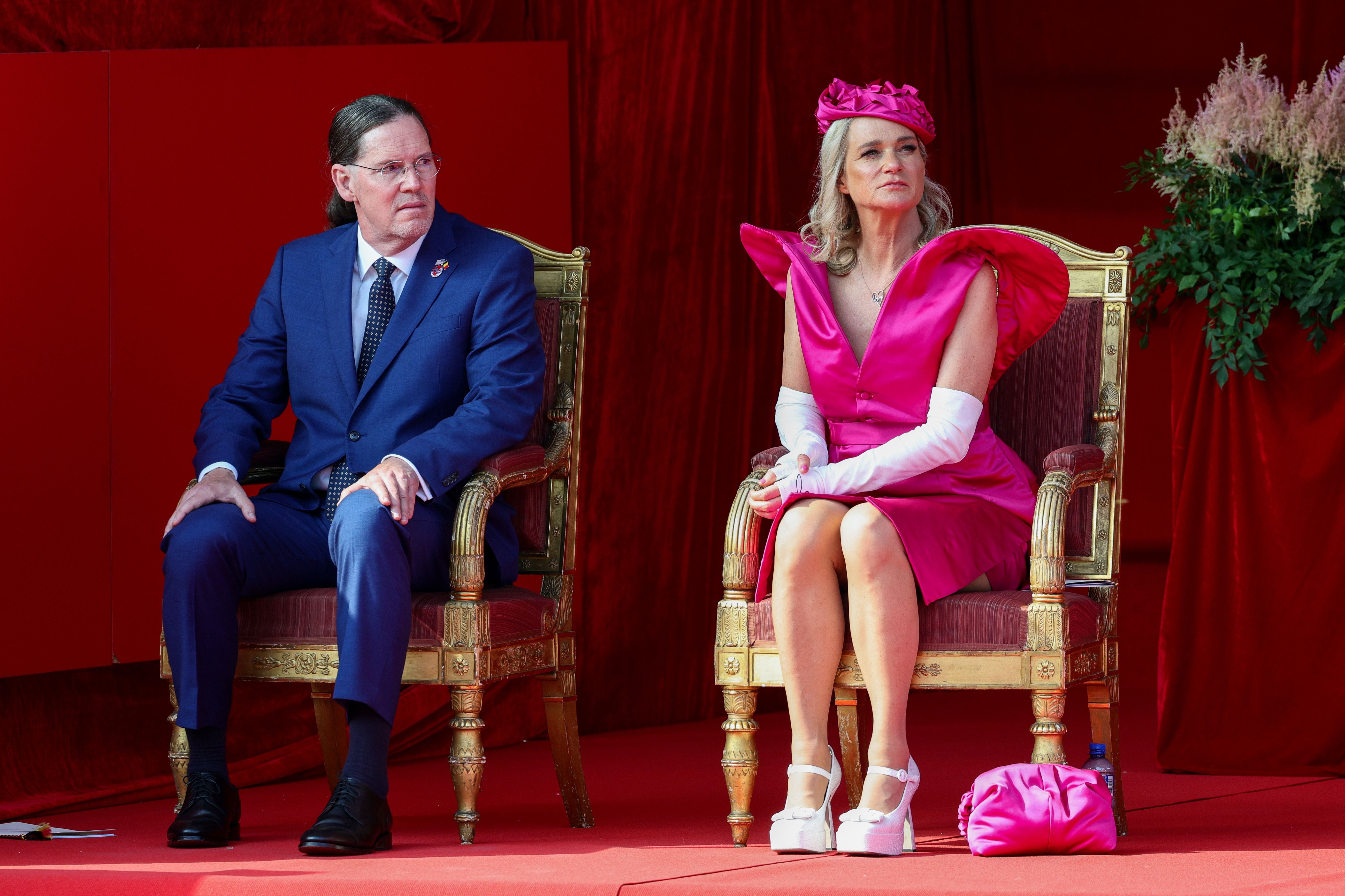 Ein Mann im Anzug und eine Frau im pinken Kleid sitzen auf prunkvollen Stühlen vor rotem Hintergrund.