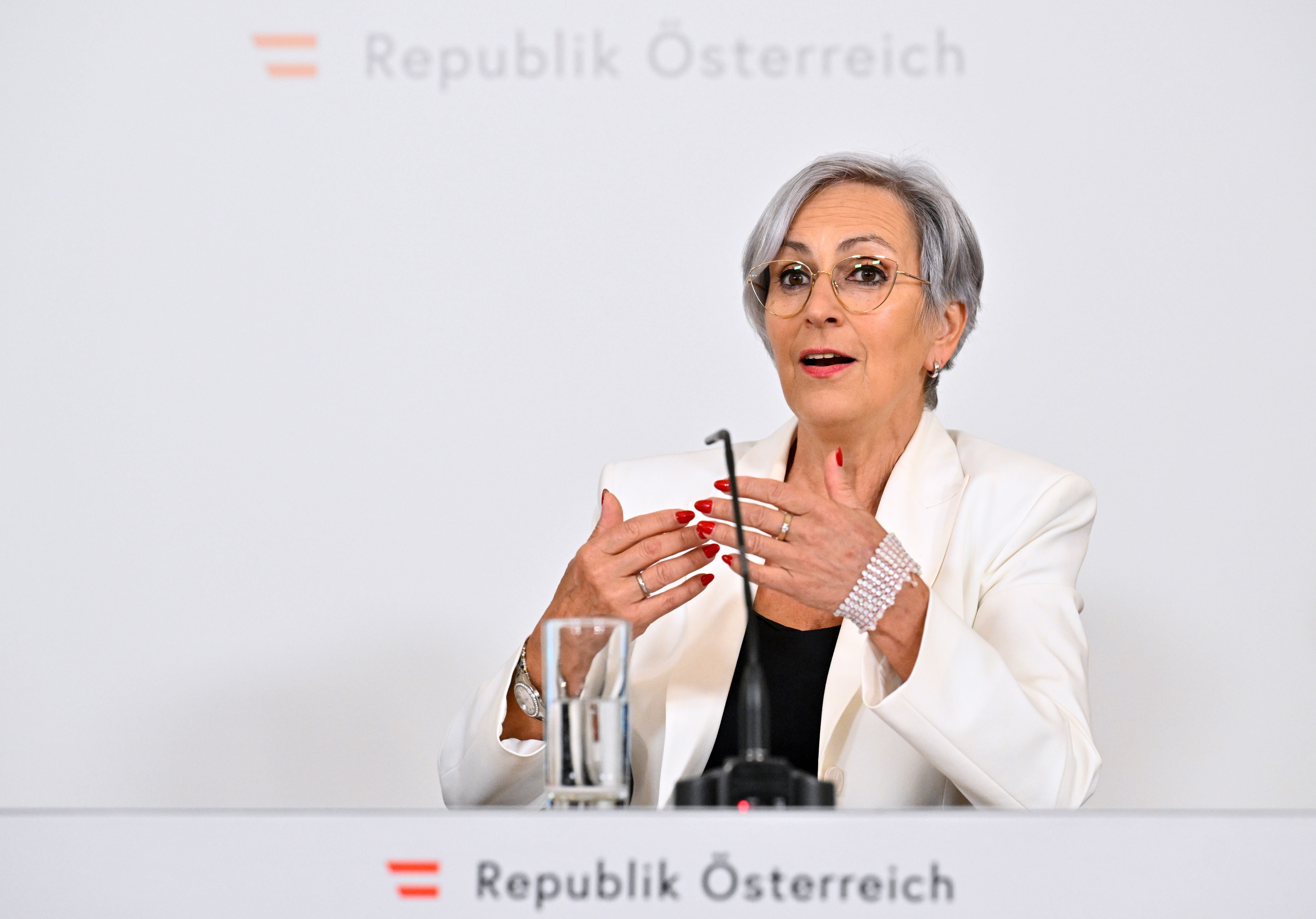 Eine Frau im weißen Blazer spricht bei einer Pressekonferenz in Wien am Mikrofon.
